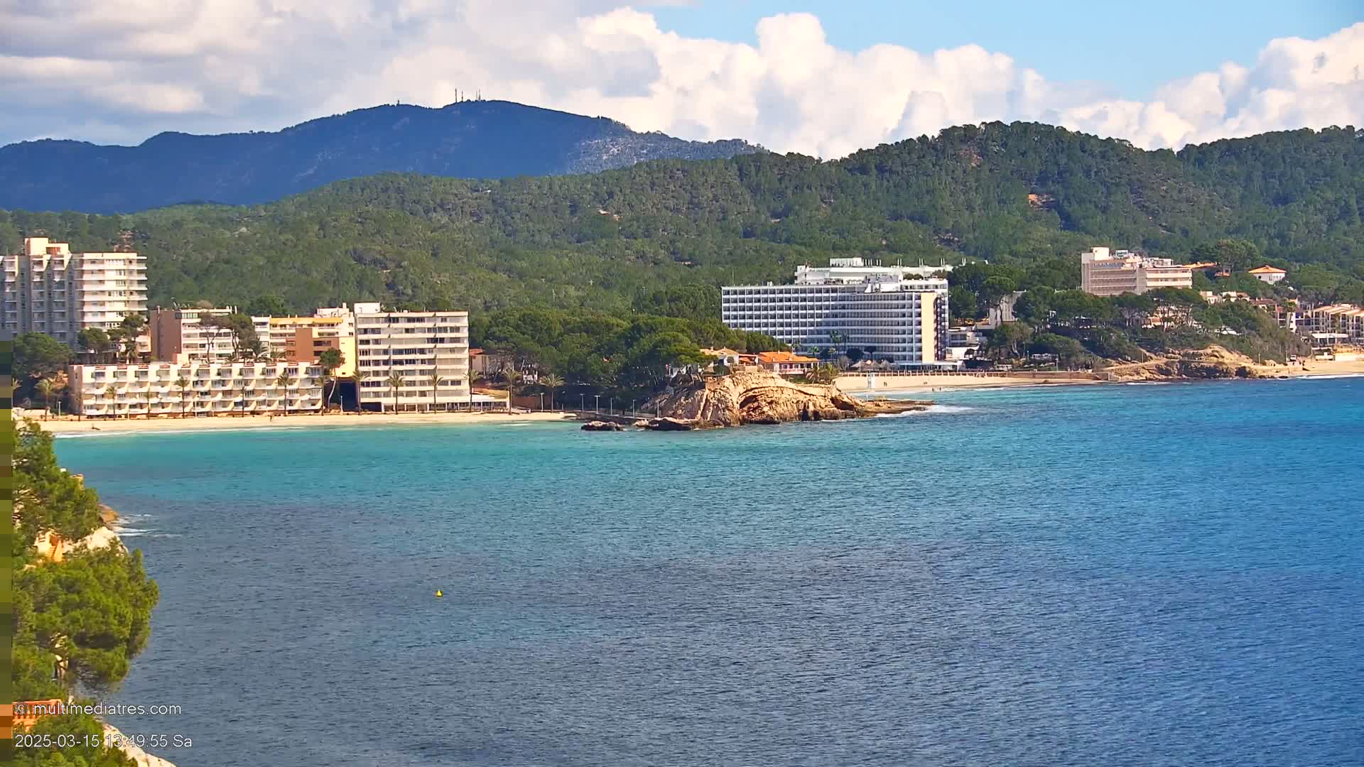 A partly cloudy day reveals a picturesque coastal scene featuring several hotels situated along a sandy beach, a rocky outcrop in the turquoise water, and verdant hills rising in the background.