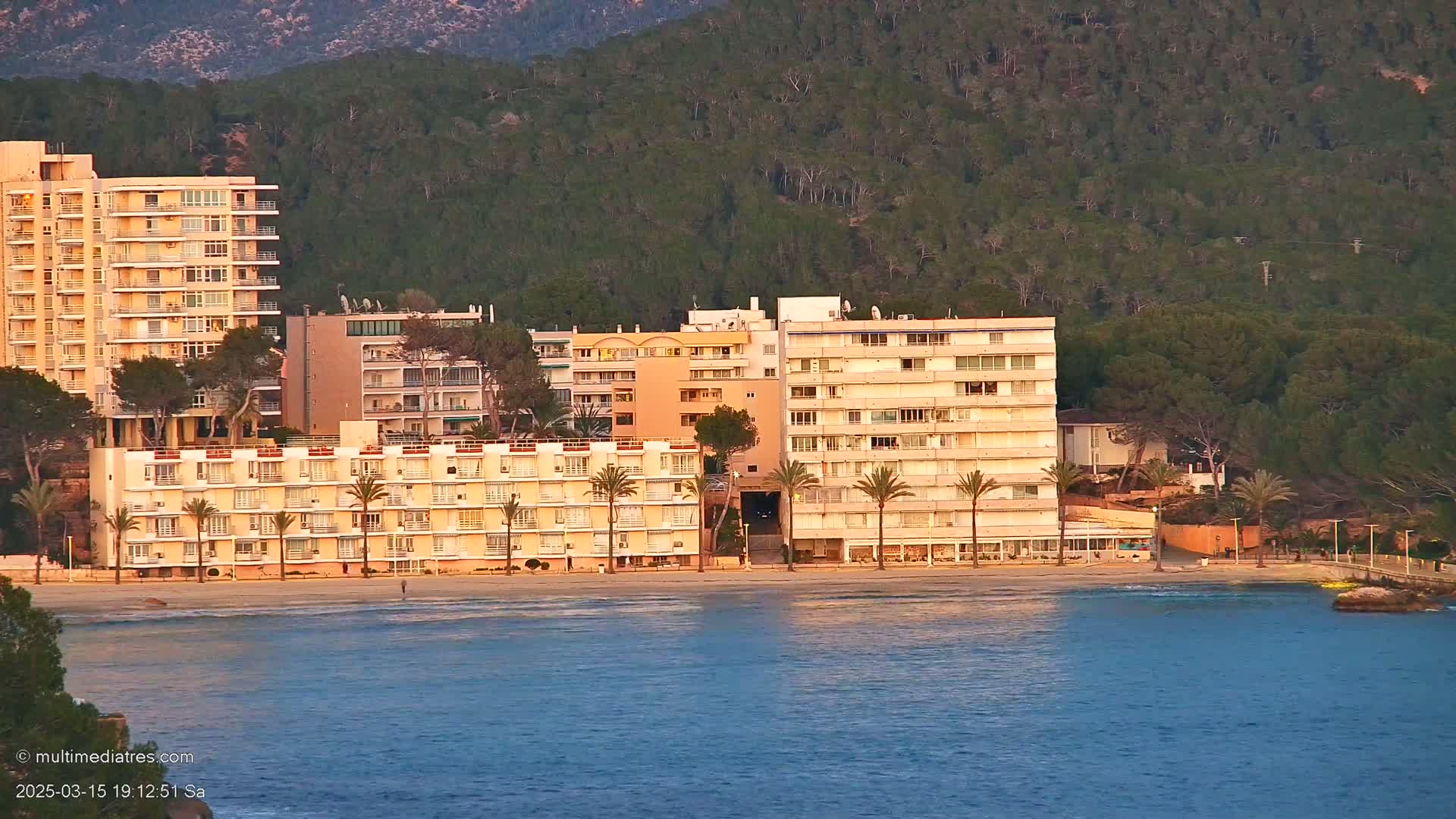 Several multi-story buildings line a beach at sunset, with calm water and a forested hillside in the background.