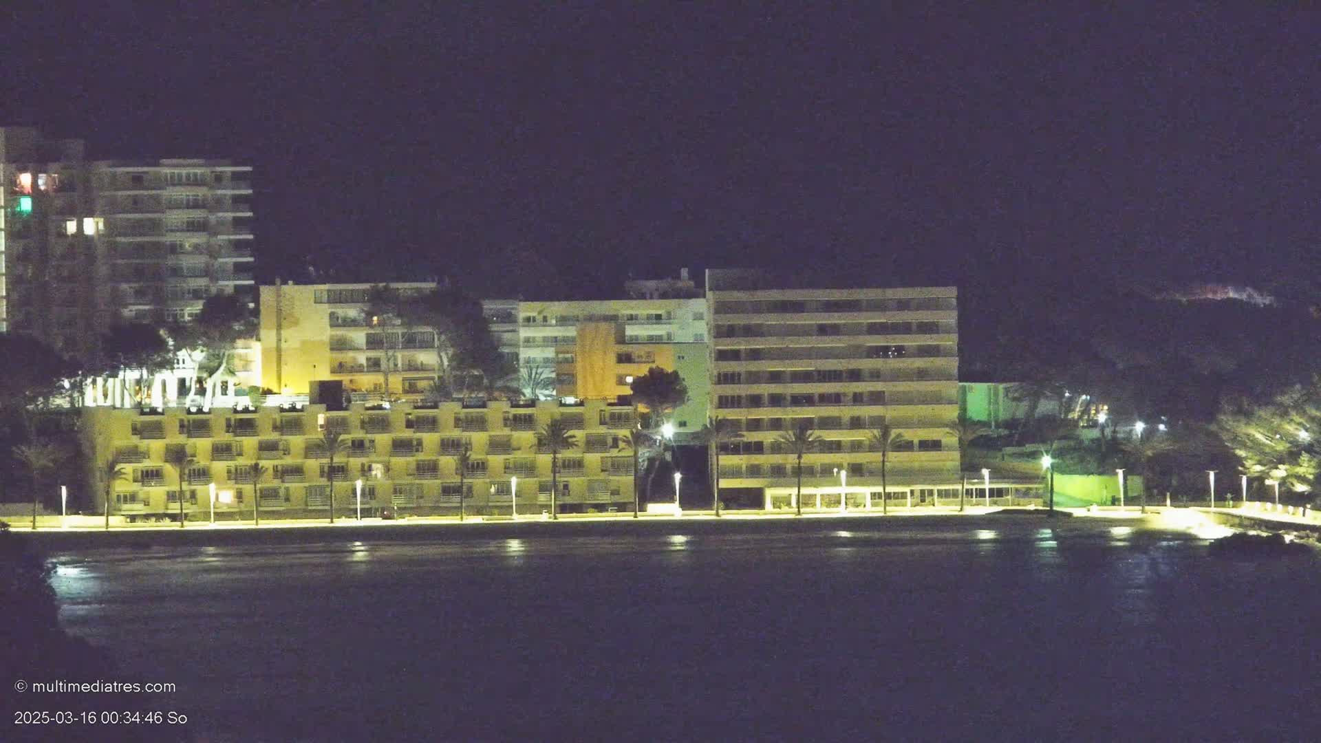A row of illuminated multi-story buildings stands along a dark waterfront at night.
