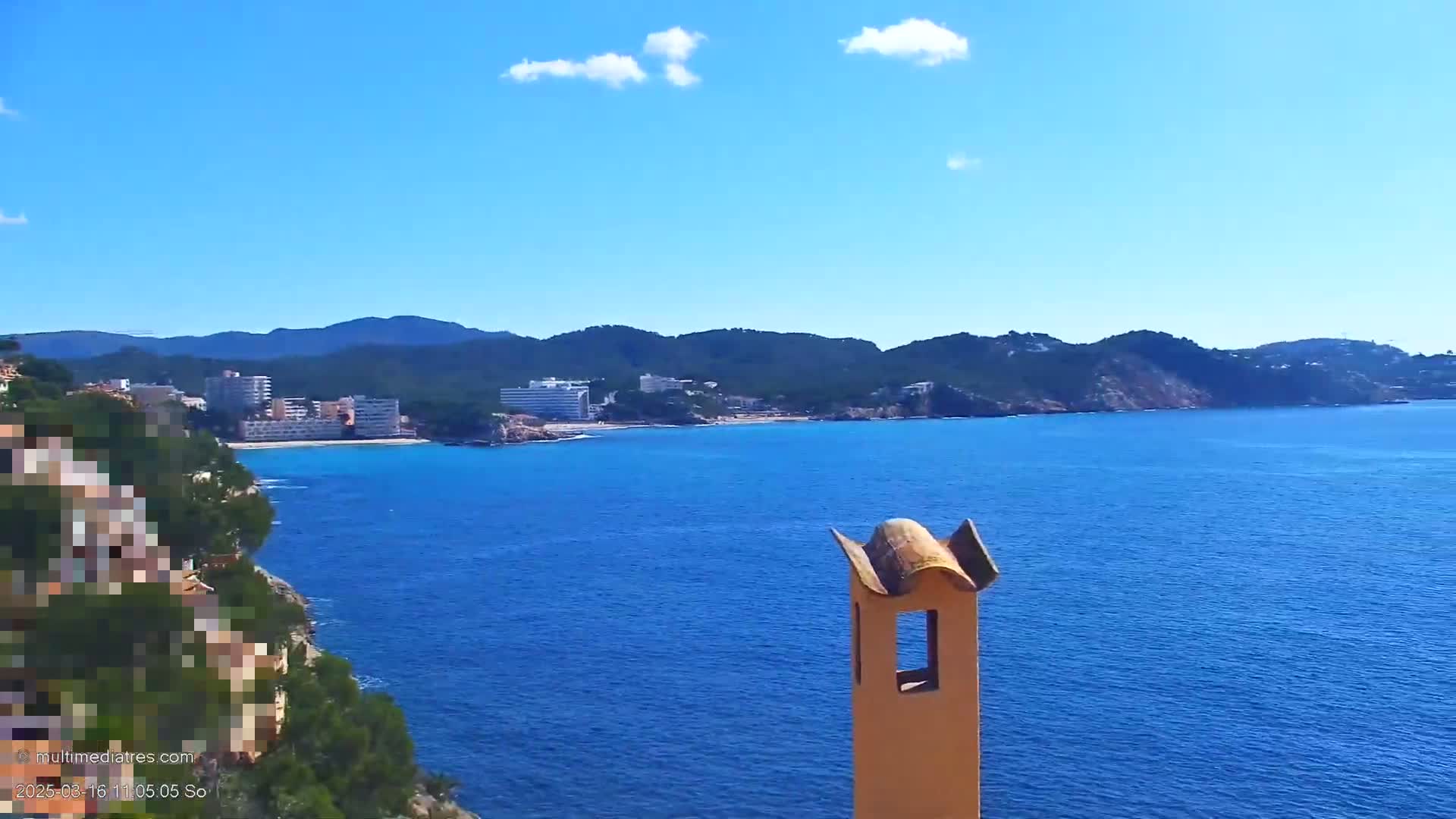 A tan chimney stands in the foreground overlooking a calm blue ocean, a coastal town, and green hills under a mostly clear blue sky.