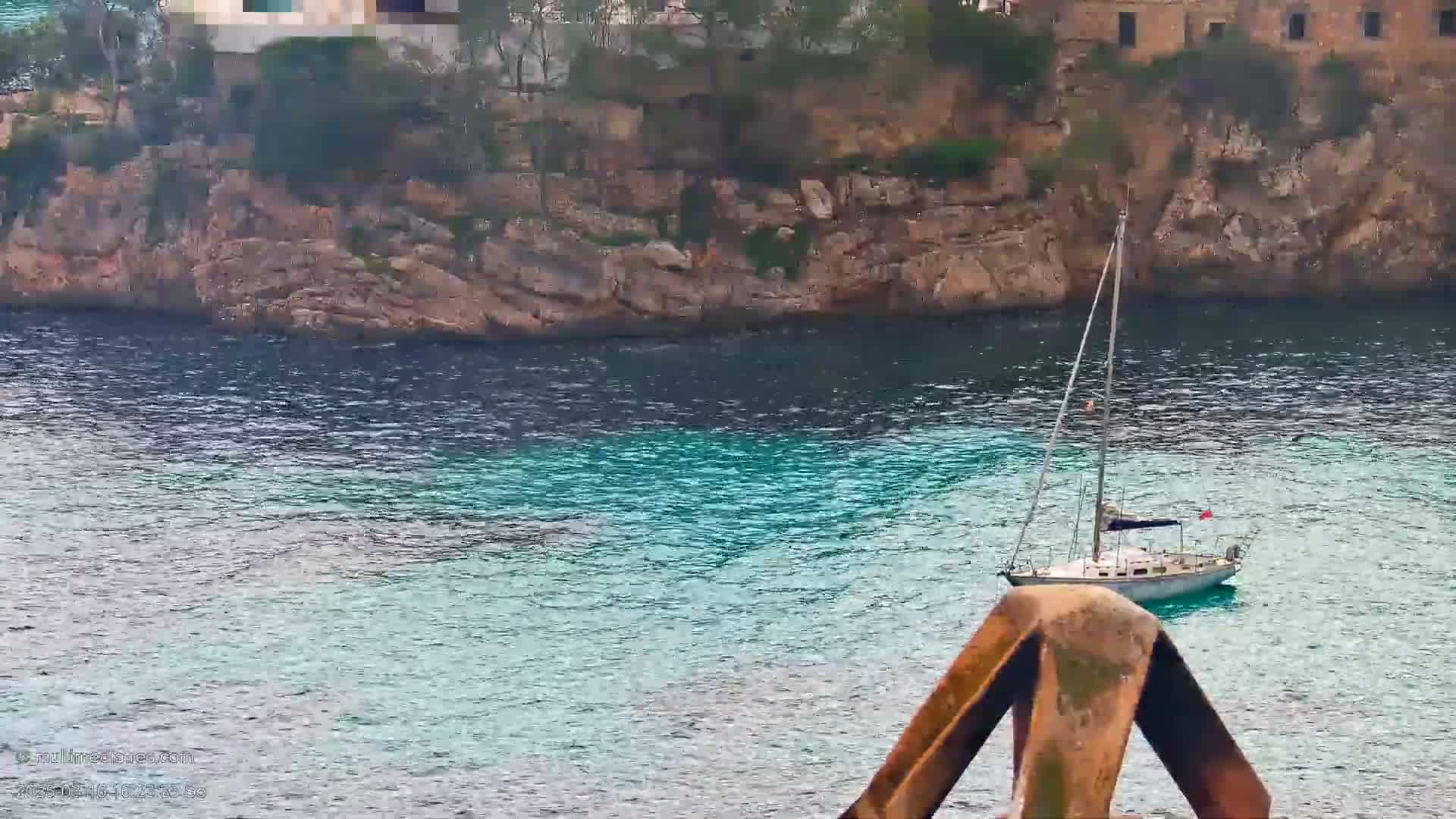A sailboat rests in calm, turquoise water near a rocky cliff overlooking buildings.