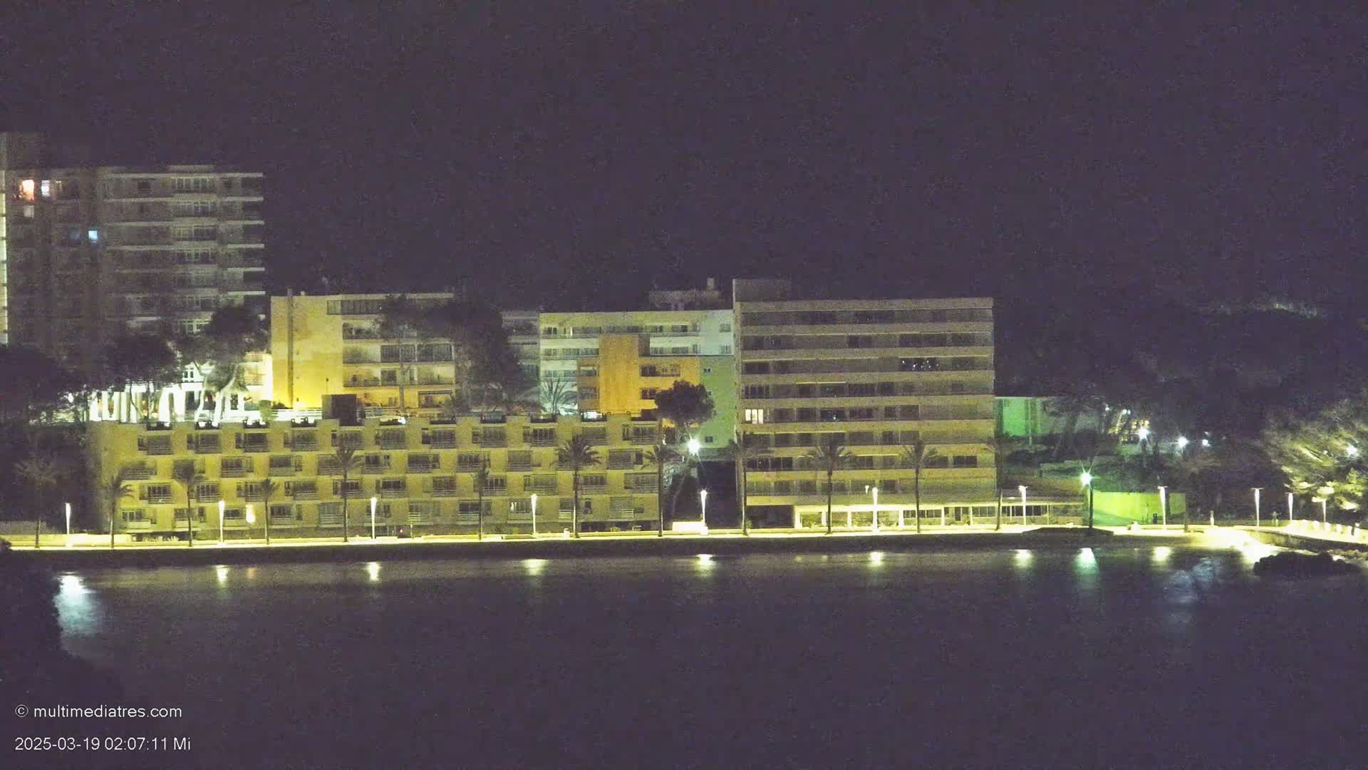 Illuminated buildings line a waterfront at night, reflected in the calm water.