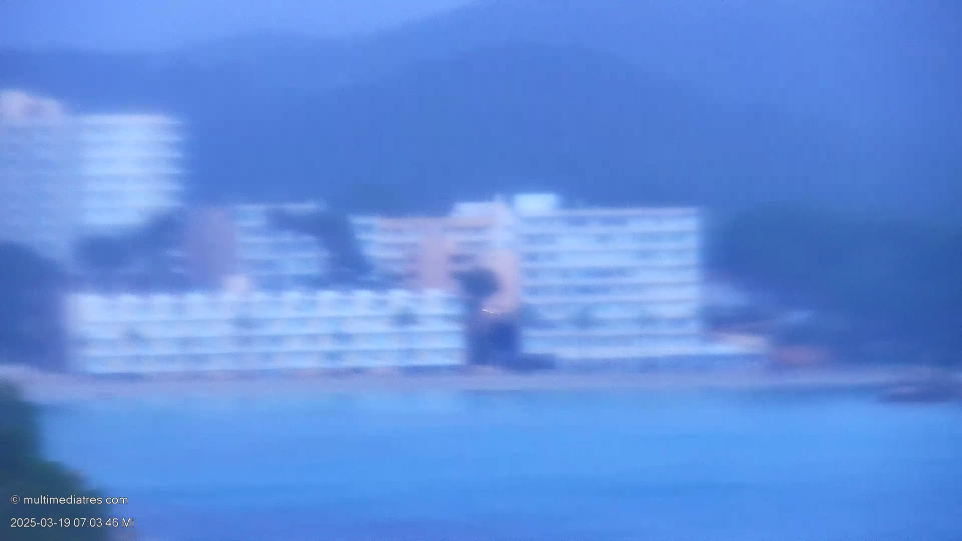 A blurry image shows a multi-story building near a body of blue water on an overcast day.