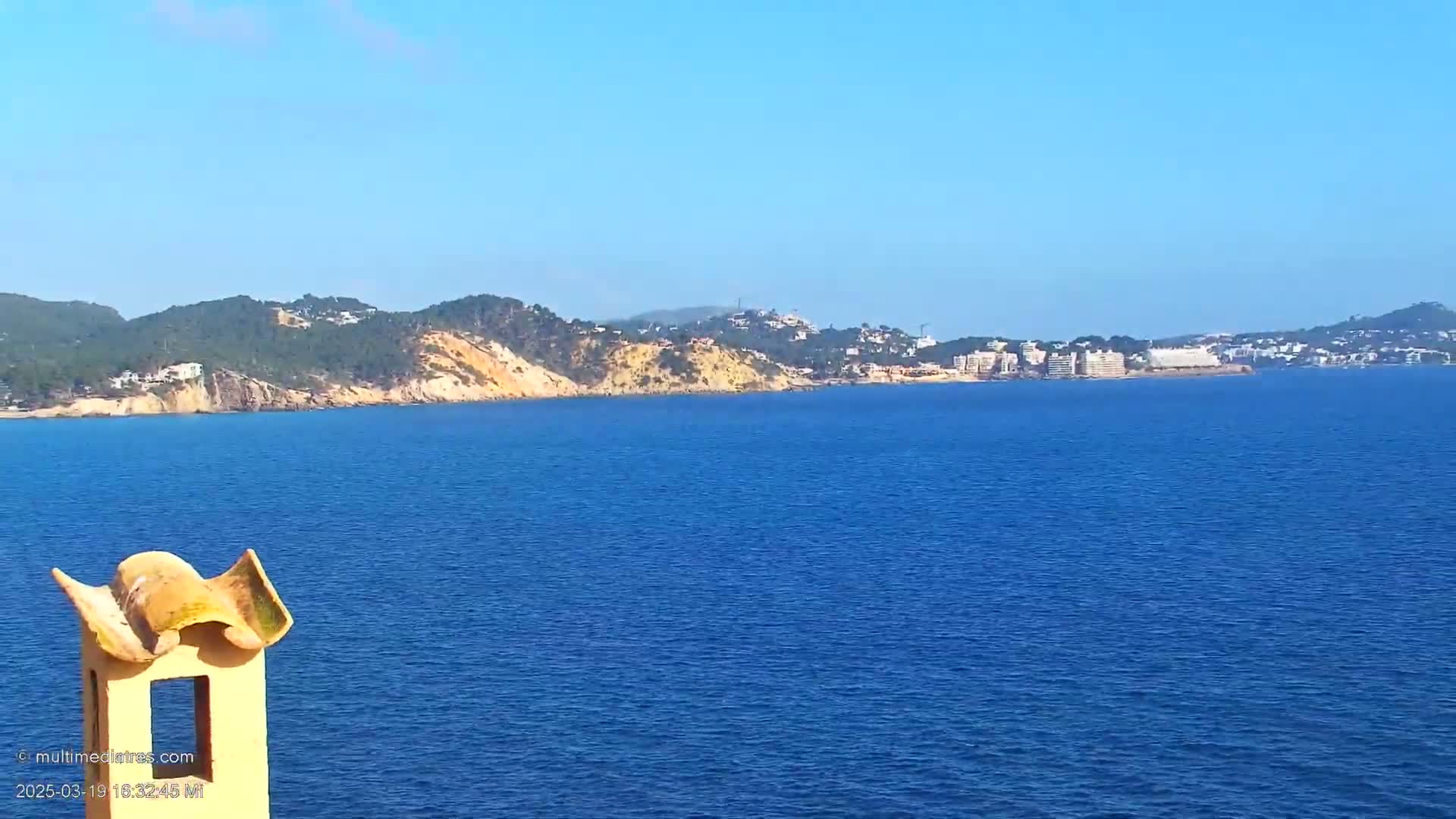 A coastal view on a clear day shows a calm blue sea extending to a distant shoreline with hills and buildings under a bright blue sky, partially obscured by a yellow-orange chimney in the foreground.