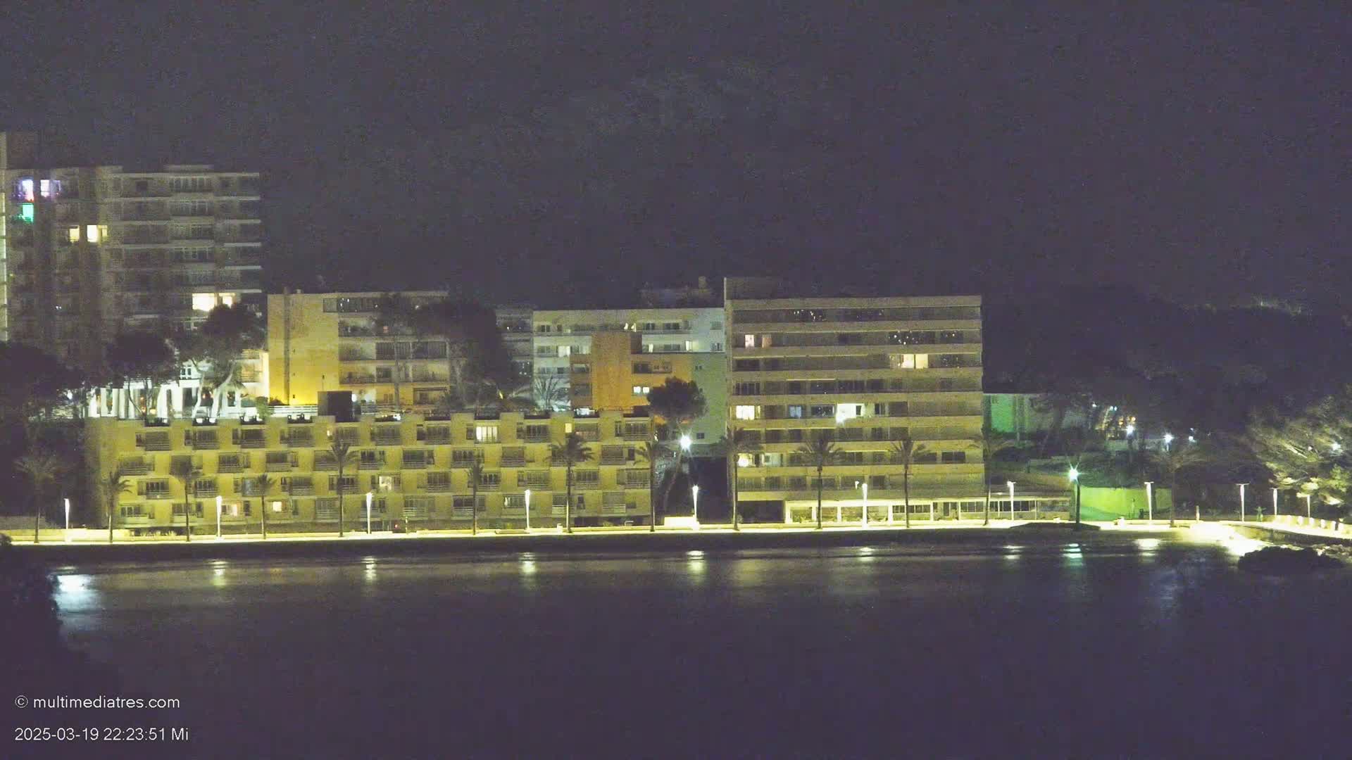 At night, several illuminated multi-story buildings line a waterfront, palm trees interspersed along a paved walkway.