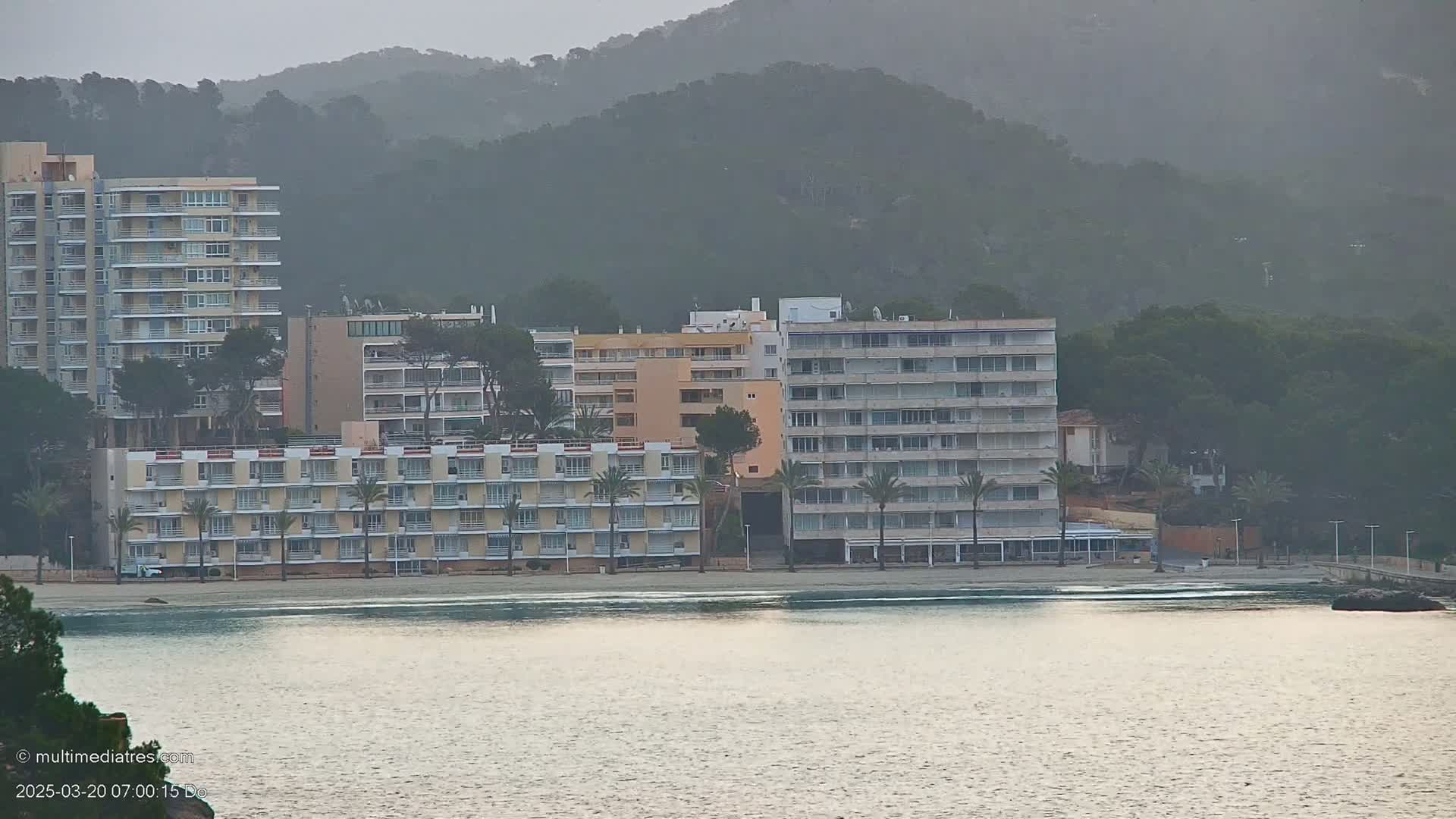 Several multi-story buildings line a beach next to calm water under an overcast sky, with a hazy mountain in the background.