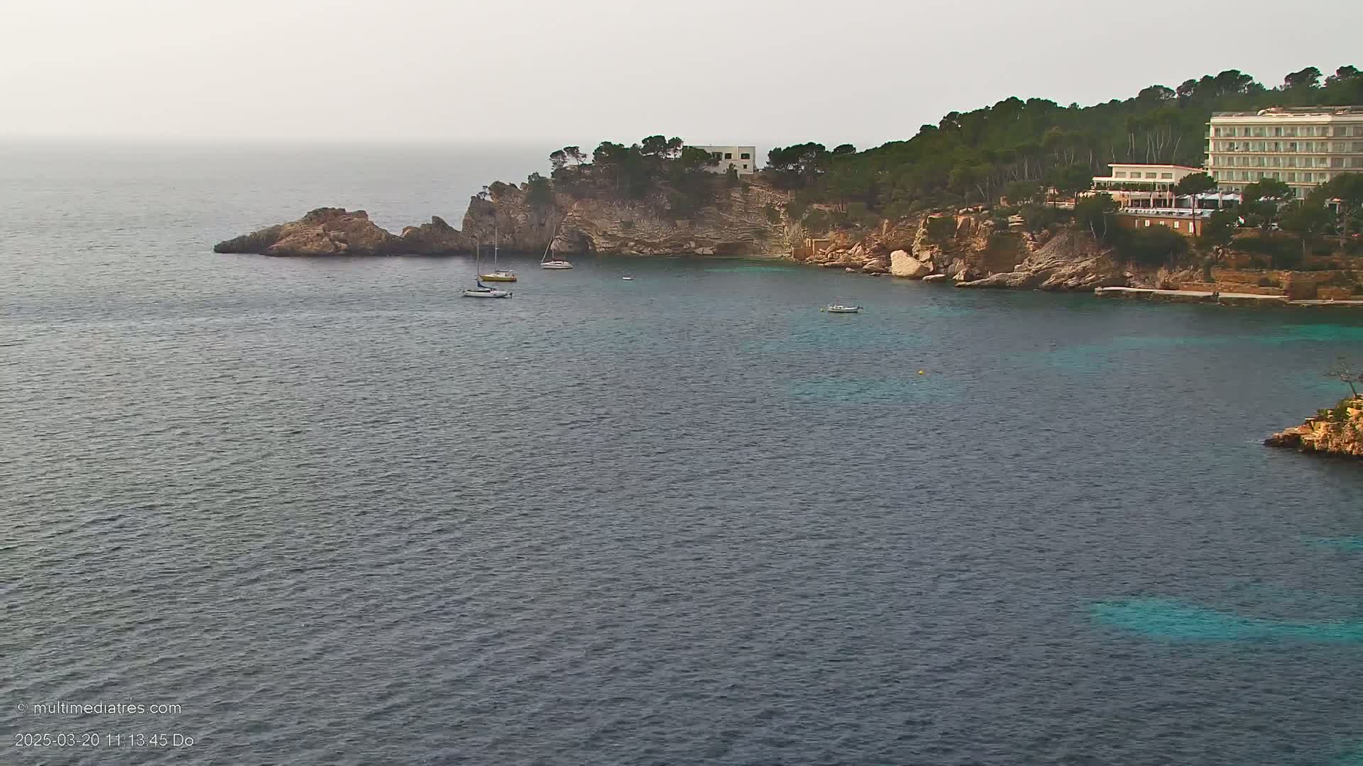 A coastal scene on a hazy day shows a calm sea with several small boats near a rocky shore, where a large hotel sits nestled among green trees.