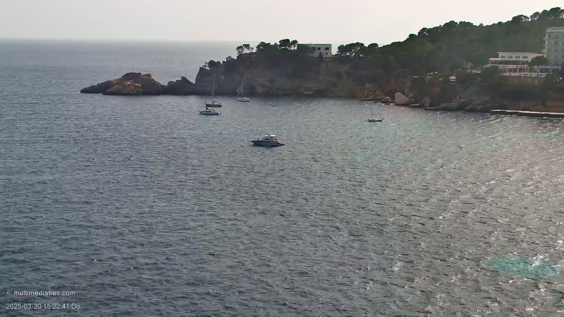 Several boats are anchored in a calm, gray sea near a rocky cliff with buildings and trees on top.