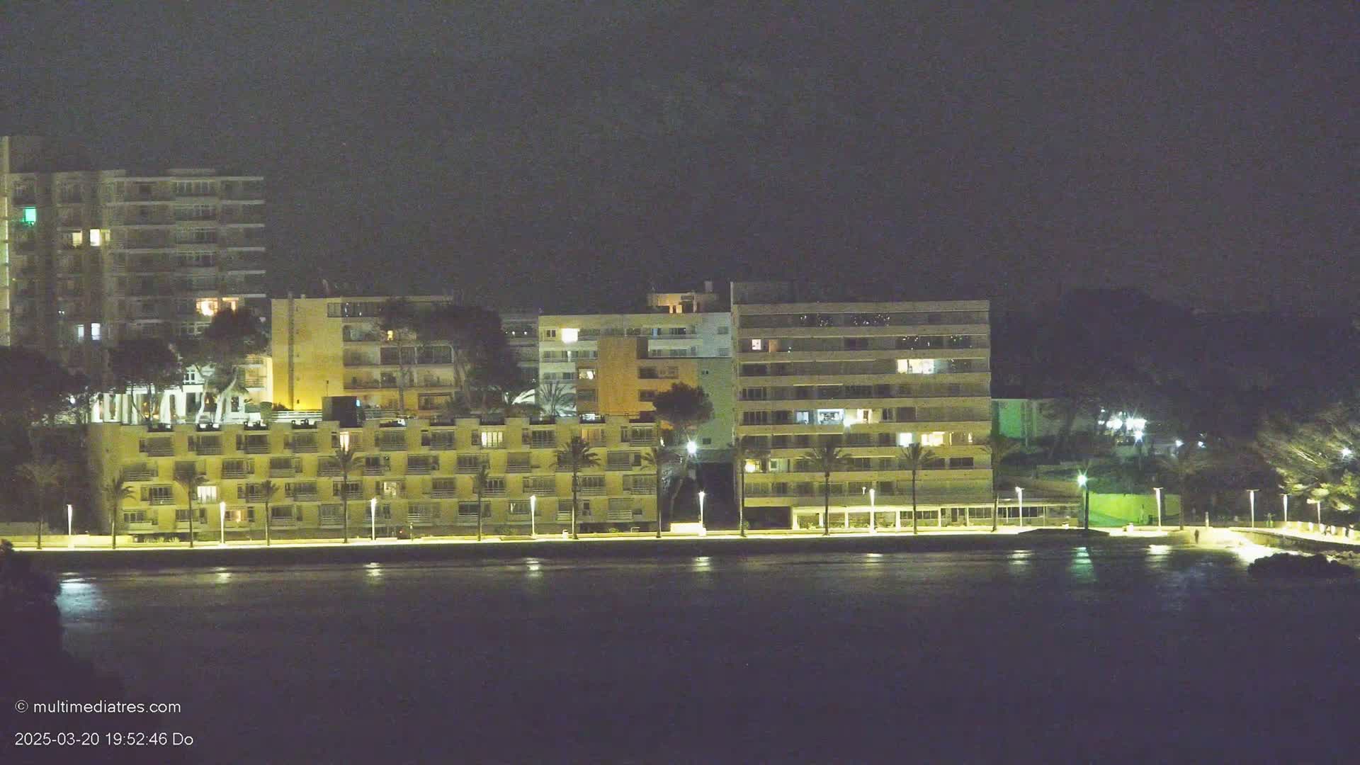 A row of lit apartment buildings stands along a dark waterfront at night.