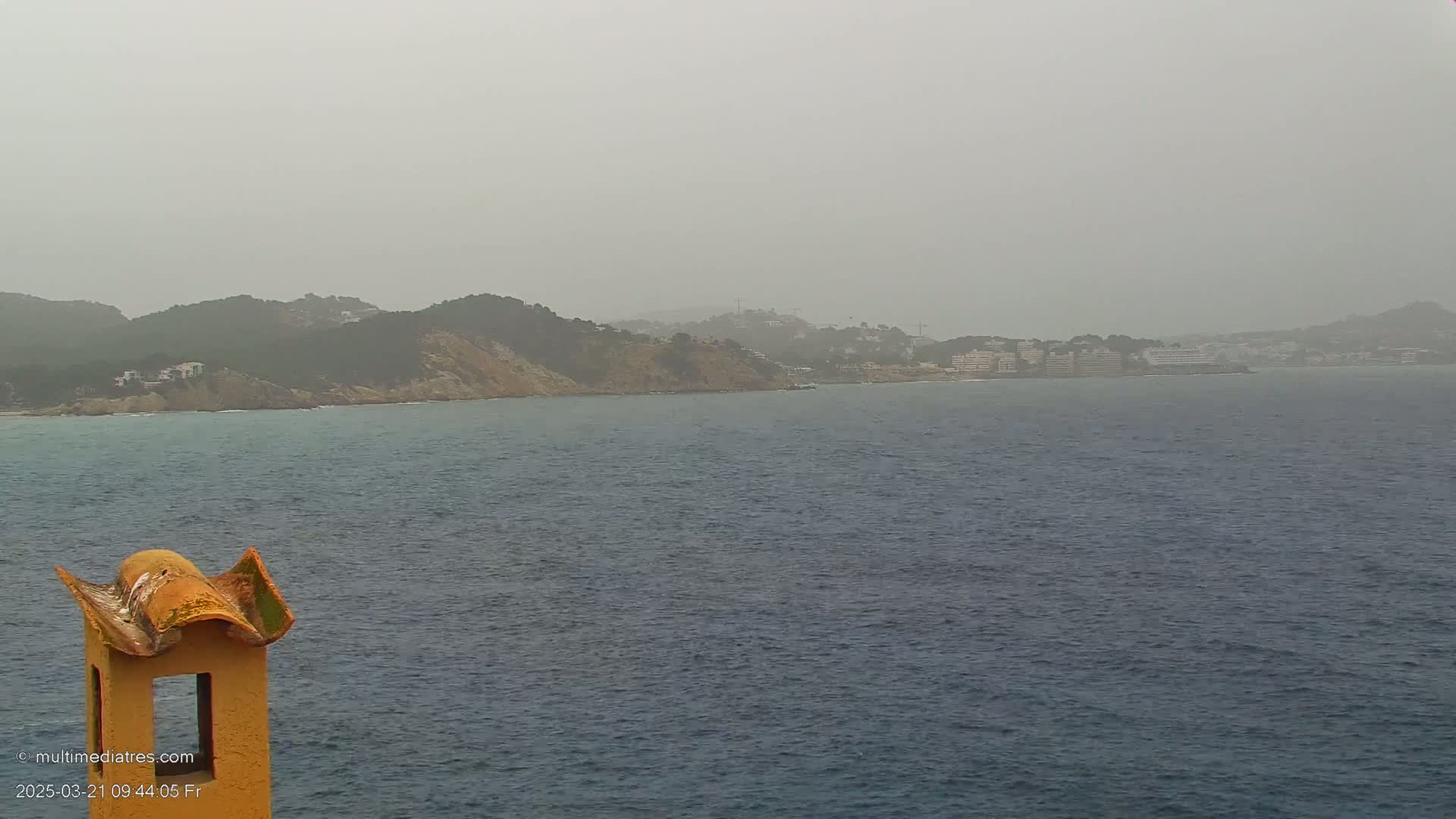 A hazy, gray day shows a calm body of water with a distant coastline featuring hills and buildings, viewed from a vantage point near an orange chimney.