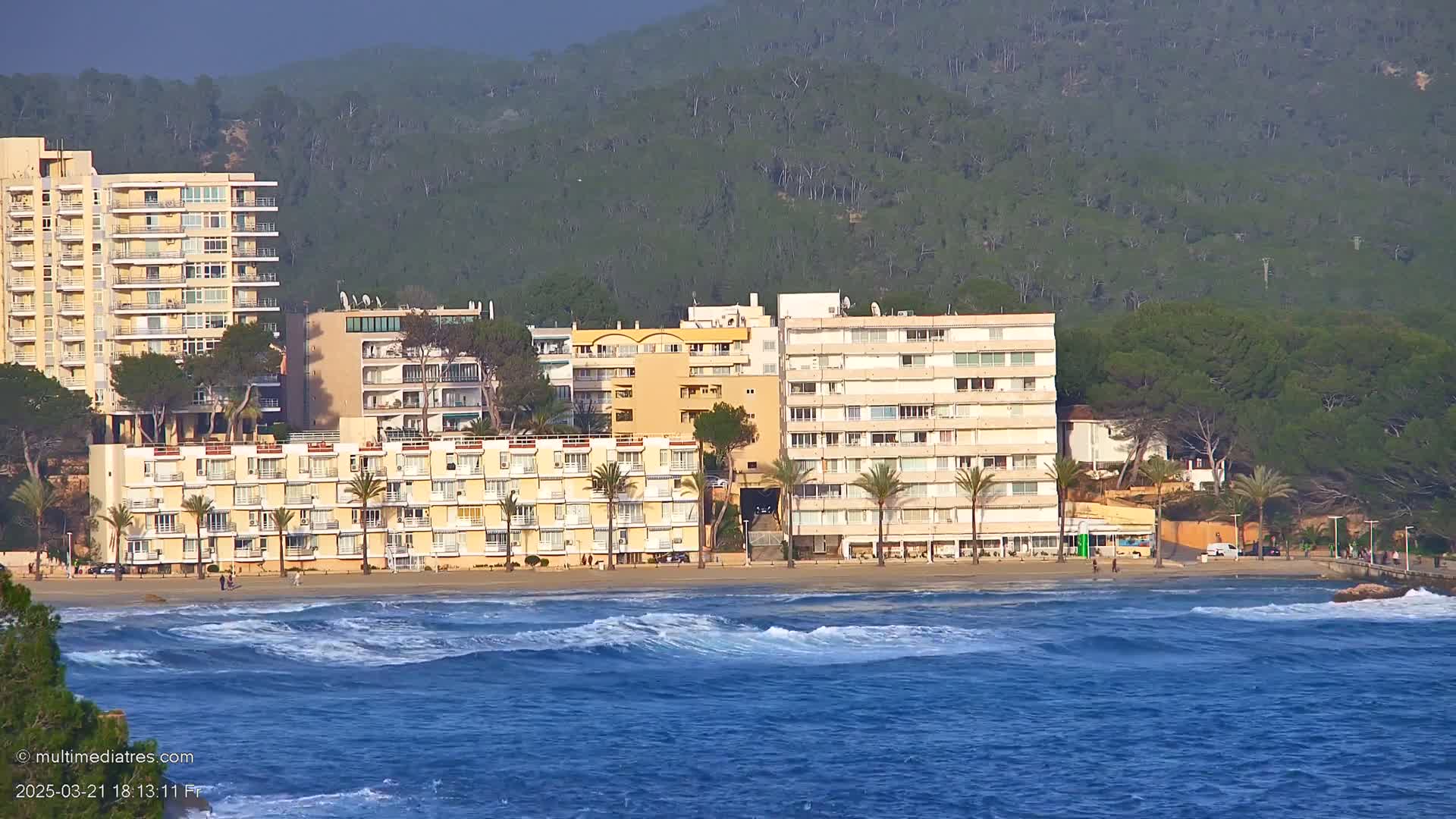 Several multi-story buildings are situated along a sandy beach, with moderate waves rolling into the ocean under a partly cloudy sky.