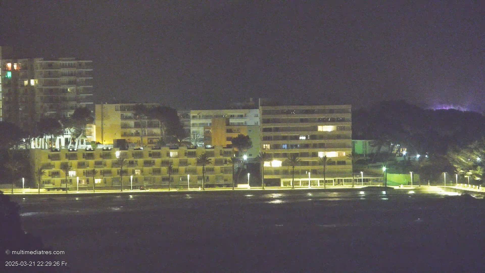 A row of lit beachfront buildings is visible at night, with dark water in the foreground.