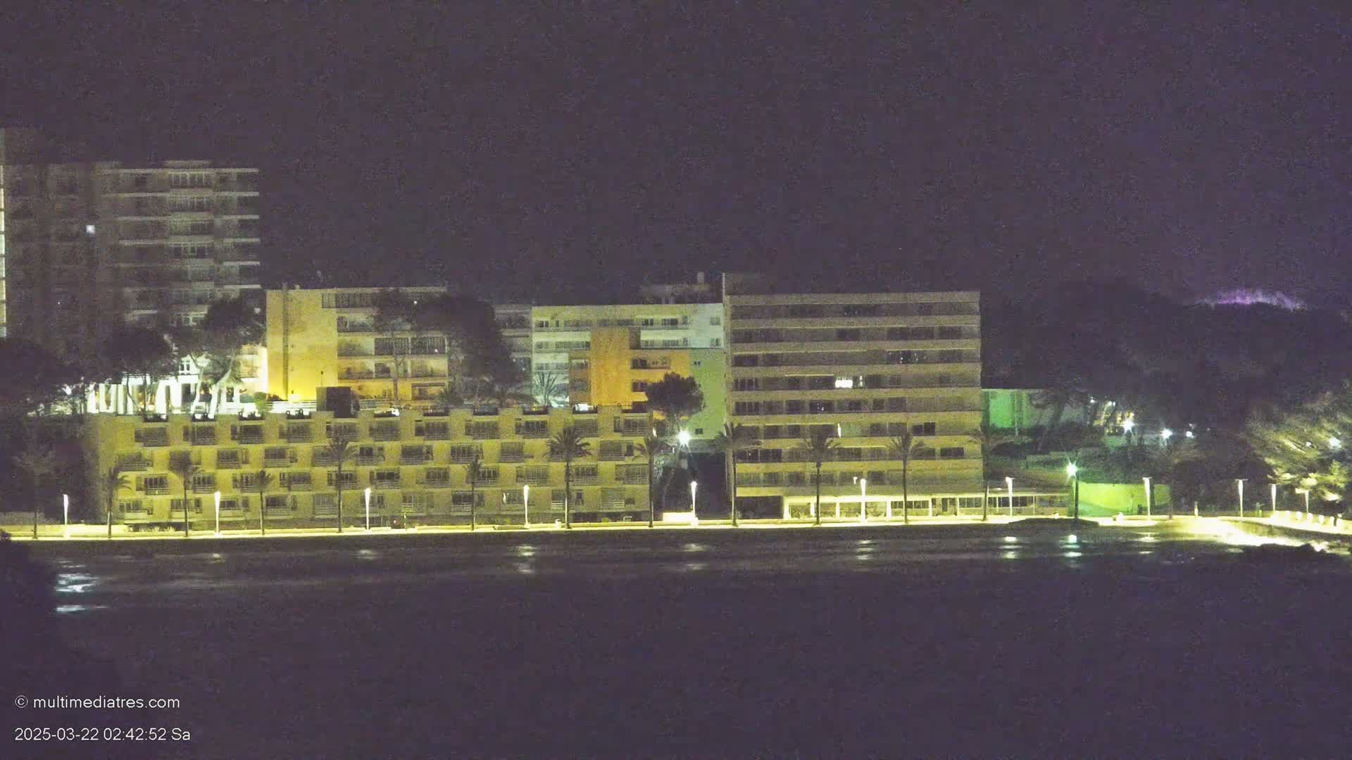 A row of illuminated apartment buildings stands along a dark shoreline at night.