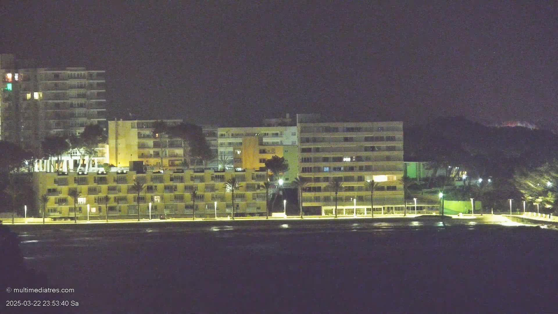 Illuminated apartment buildings line a dark shoreline at night.