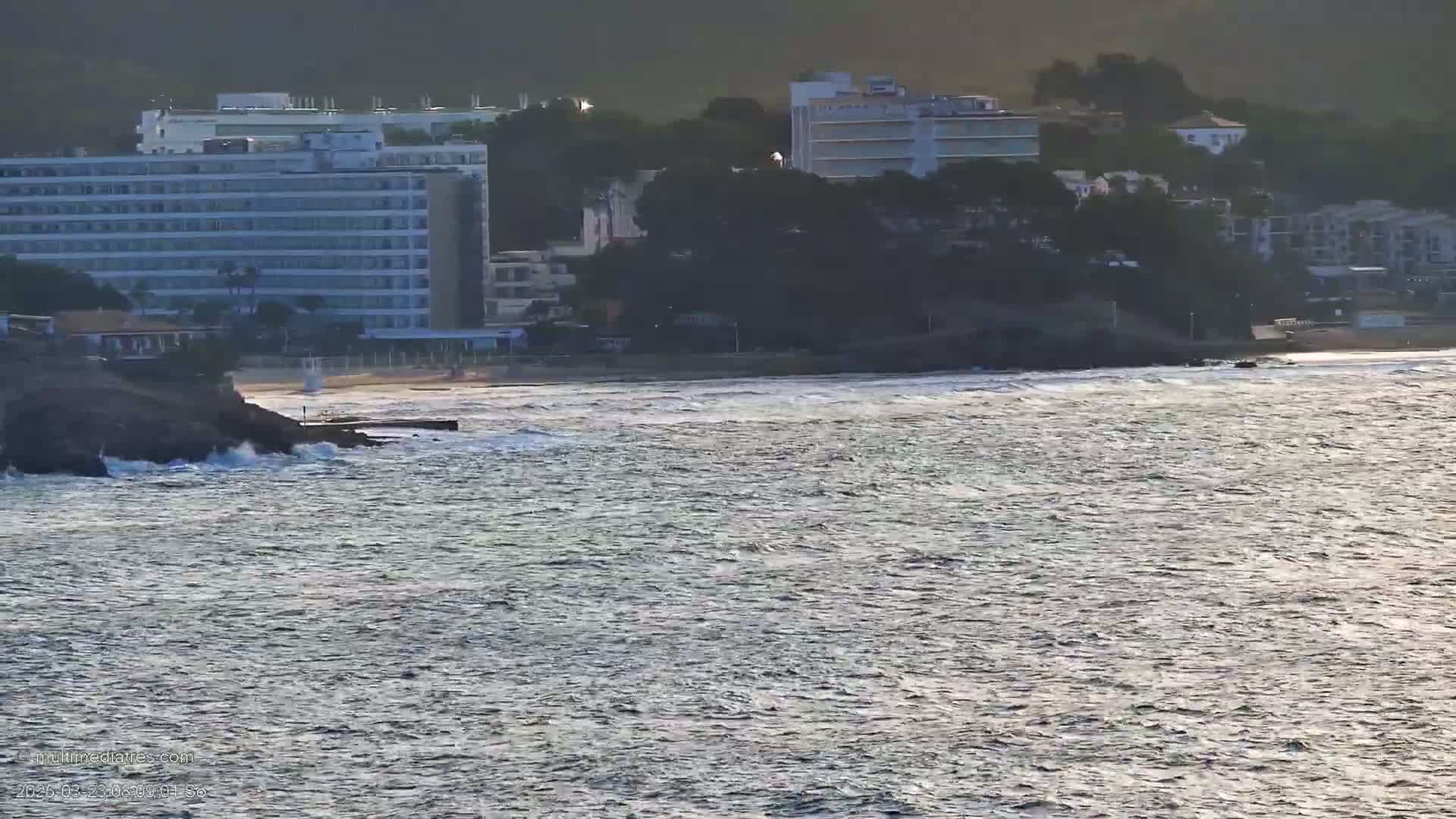 A sandy beach with a large hotel and other buildings behind it is visible from across a choppy sea.
