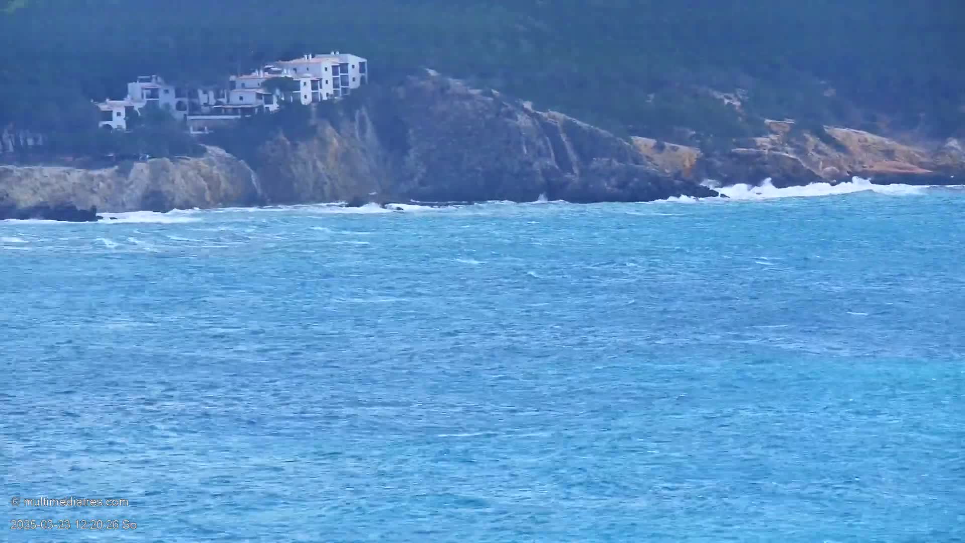 White buildings sit atop a rocky cliff overlooking a choppy blue sea with small waves.
