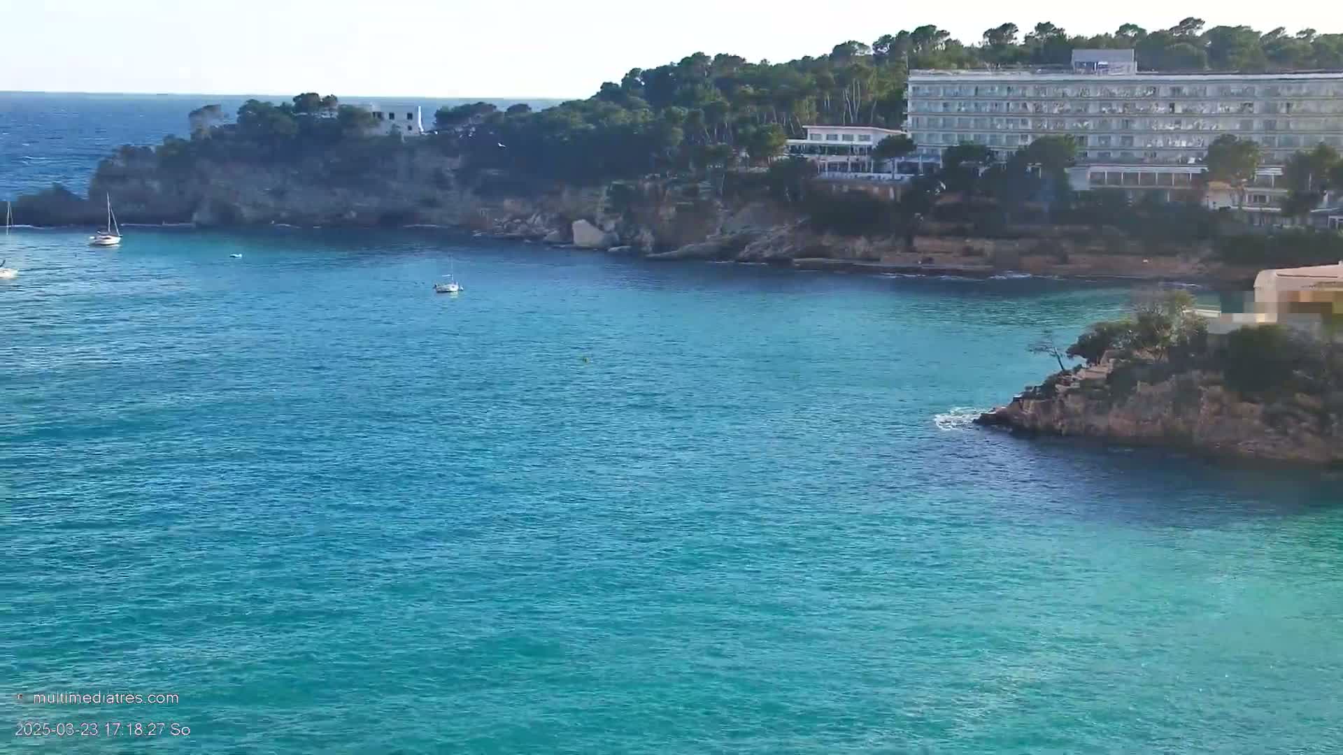 A calm, clear turquoise ocean cove is nestled between rocky shores, with a large hotel and several small boats visible.