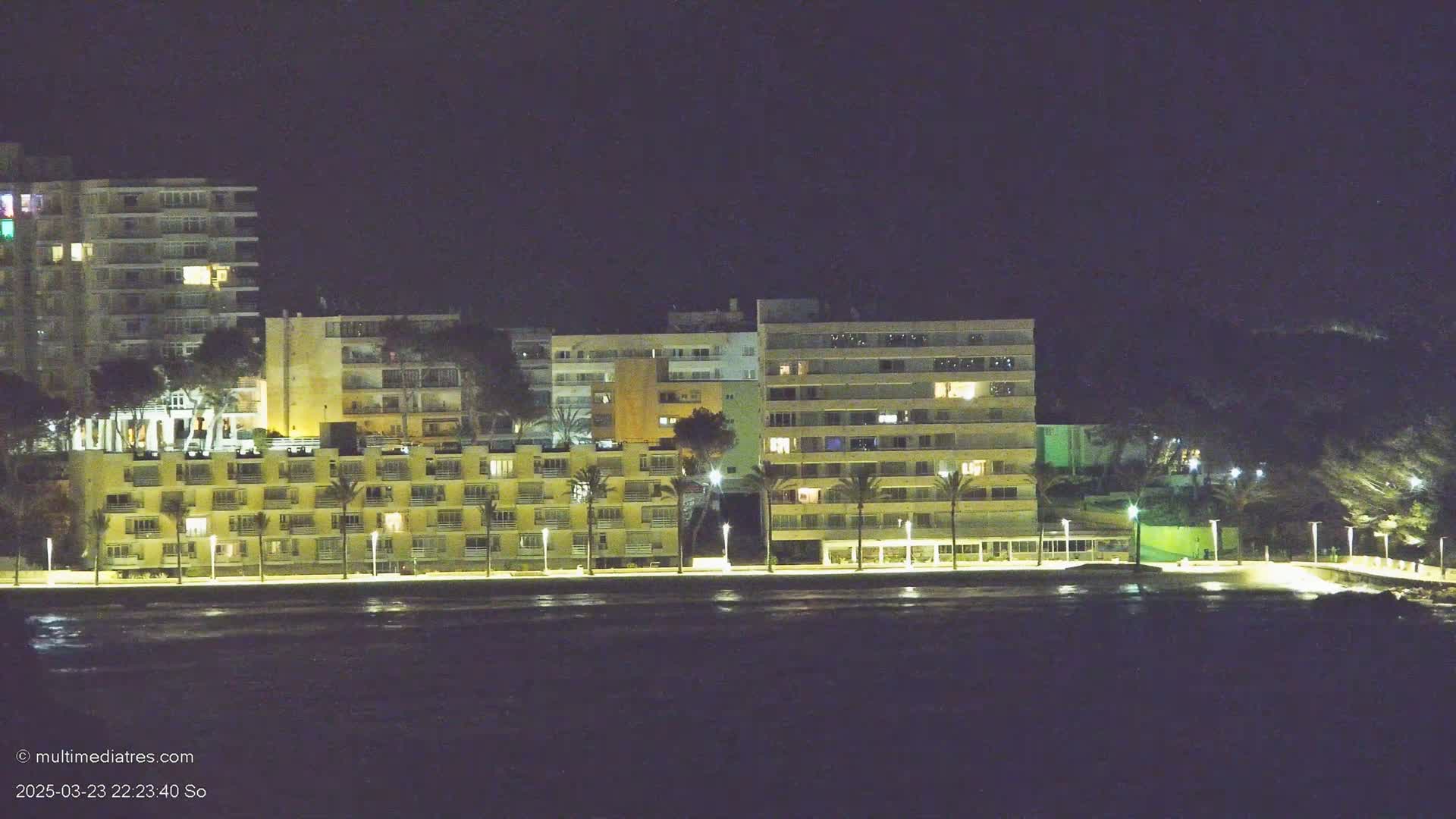 A row of illuminated apartment buildings lines a dark, calm waterfront at night.