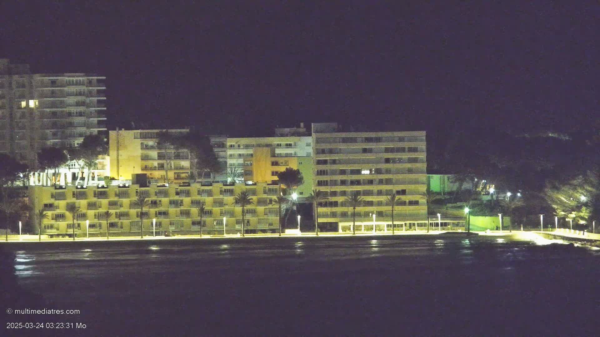 A row of illuminated beachfront buildings at night, viewed across a dark body of water.