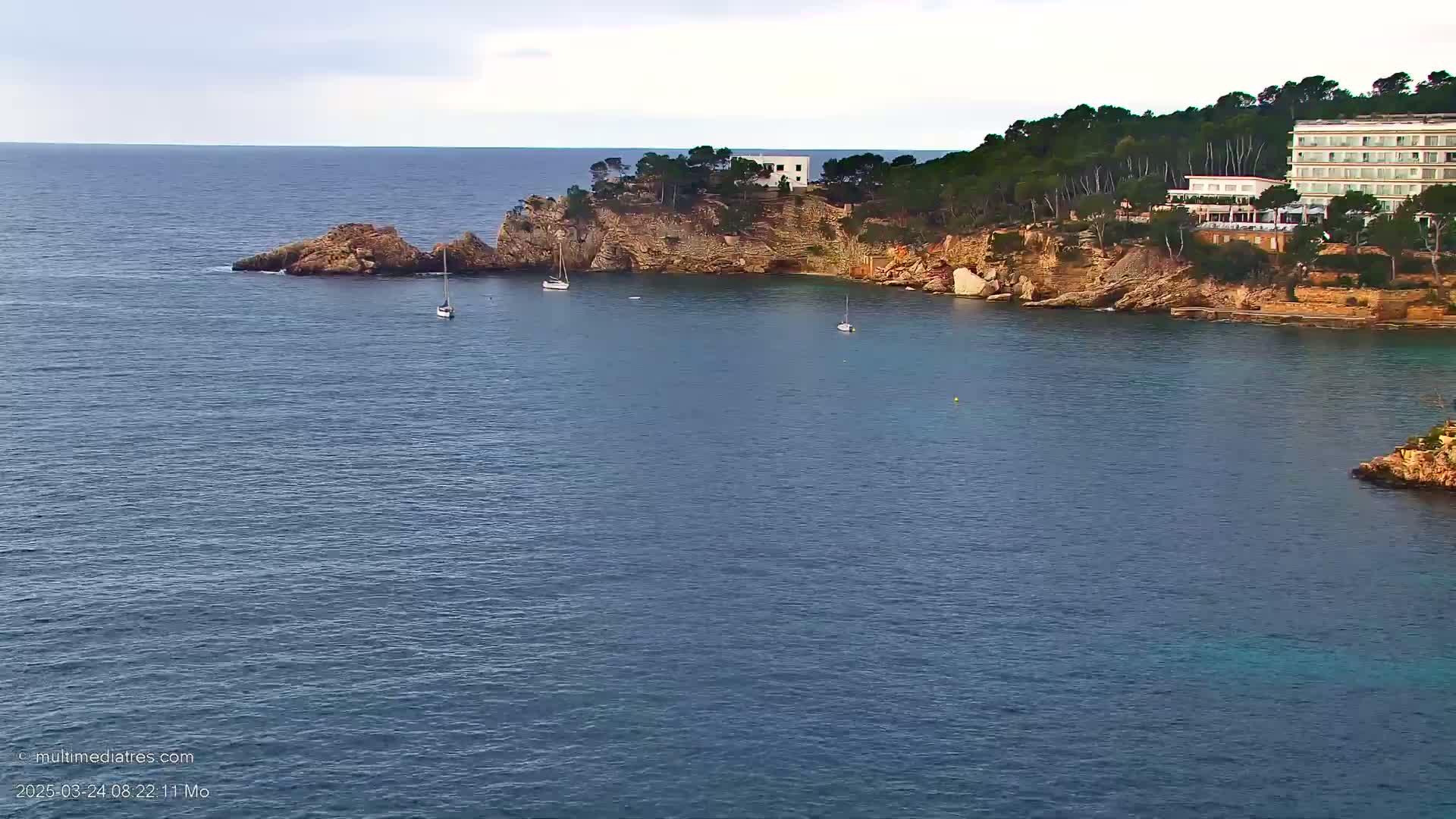 A calm, blue sea laps against a rocky coastline, where a few sailboats are anchored near a hotel nestled amongst pine trees.