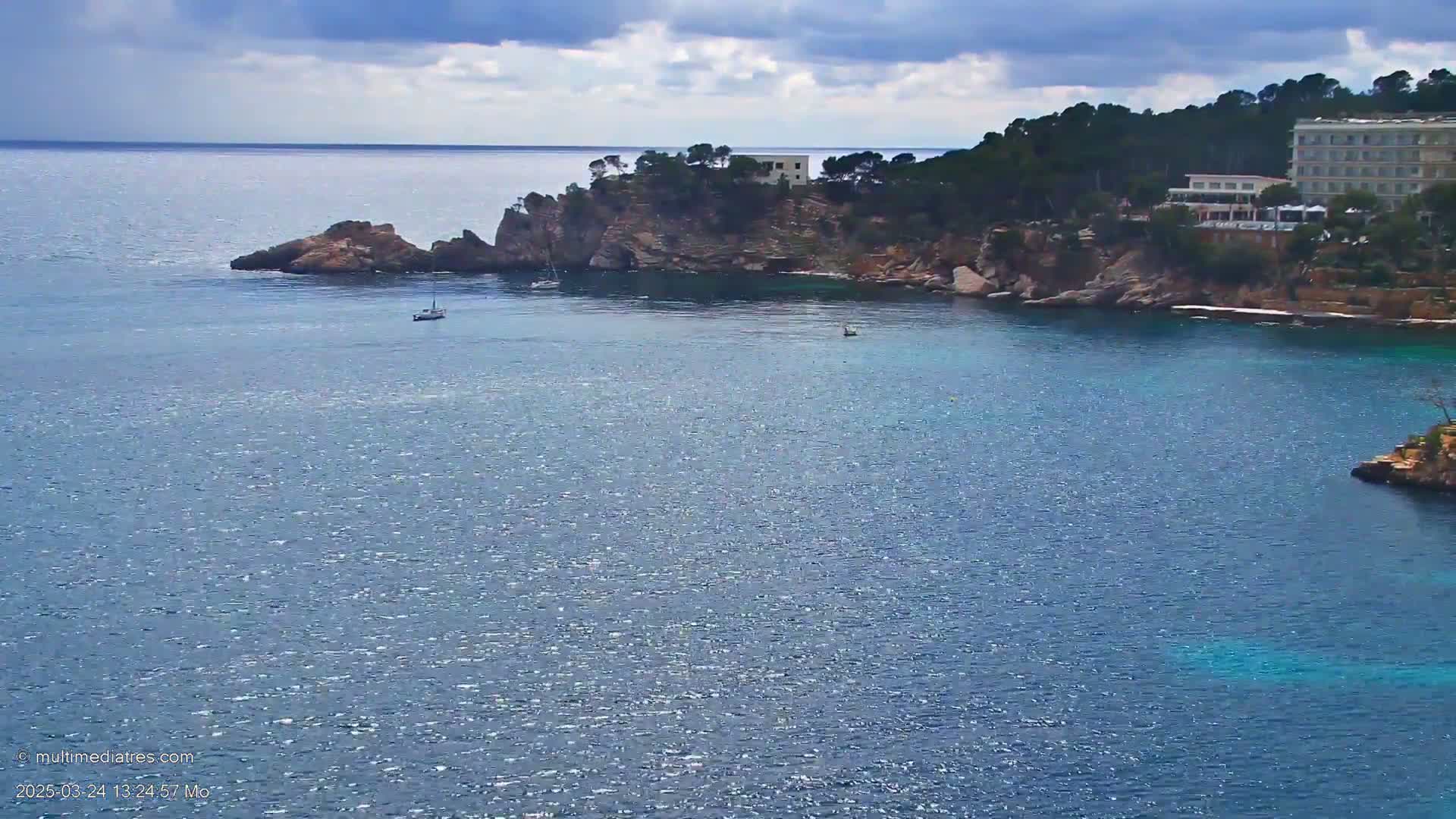 A calm, blue sea laps against a rocky coastline, where a few boats are visible, and a large building is nestled among the trees atop a cliff.