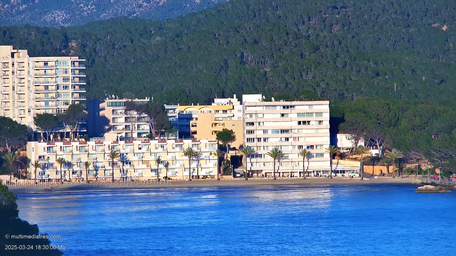 A row of multi-story buildings sits on a sandy beach next to a calm, blue ocean, with a lush green hillside rising behind them.