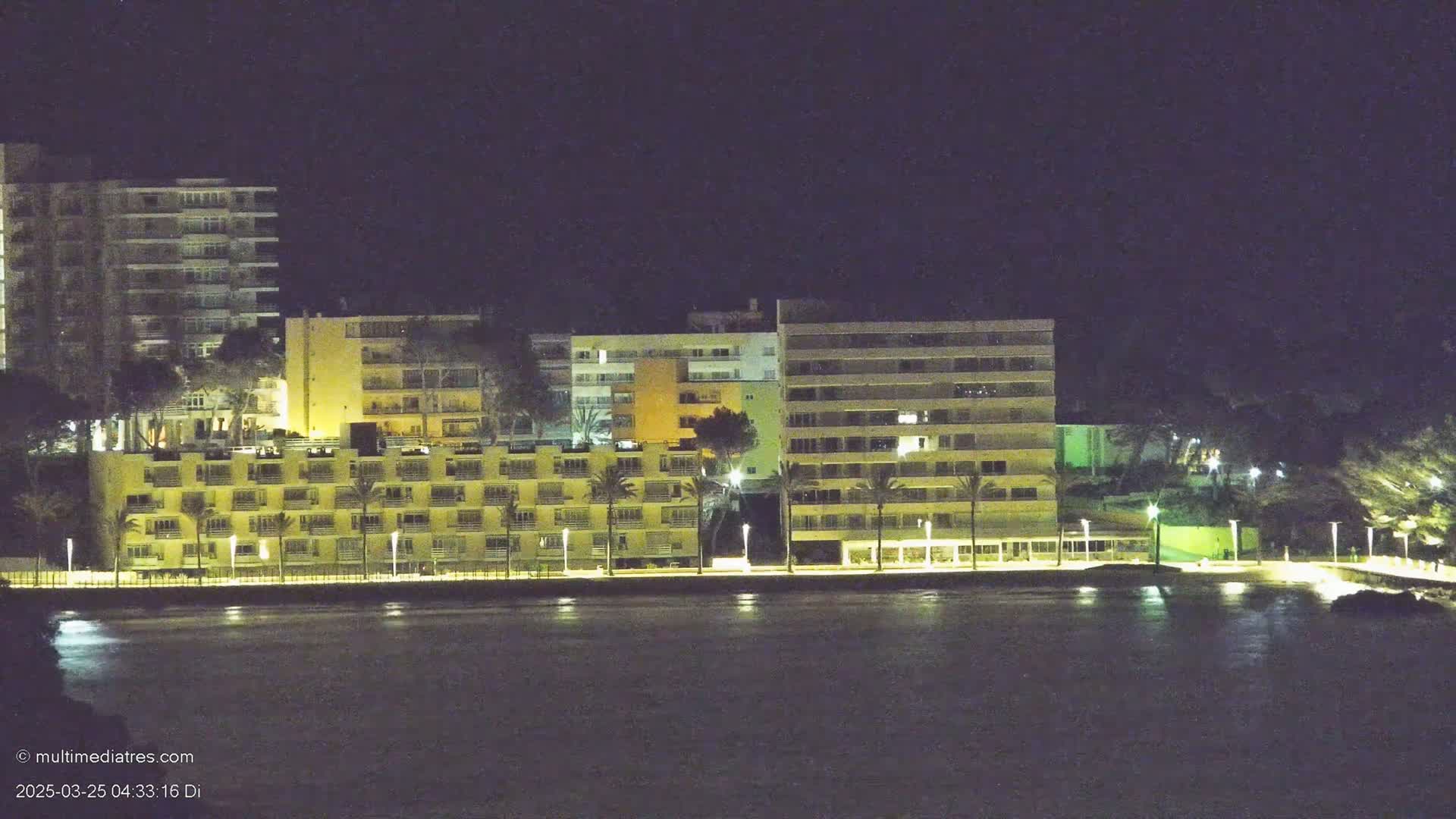 A row of brightly lit multi-story buildings stands along a dark waterfront at night.