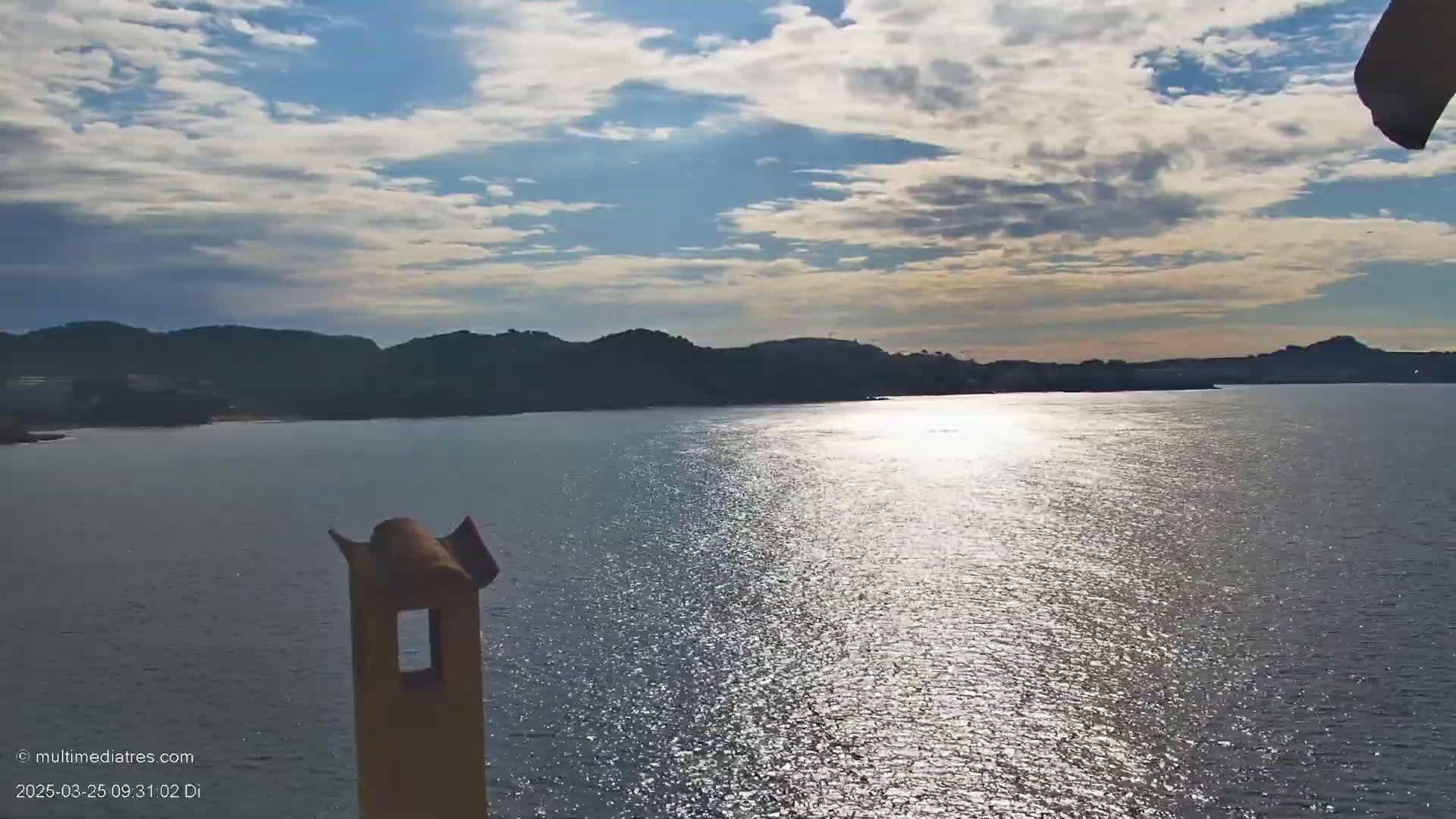 A partly cloudy day shows a calm body of water reflecting sunlight, with dark hills and a coastline in the background, partially obscured by a chimney-like structure in the foreground.
