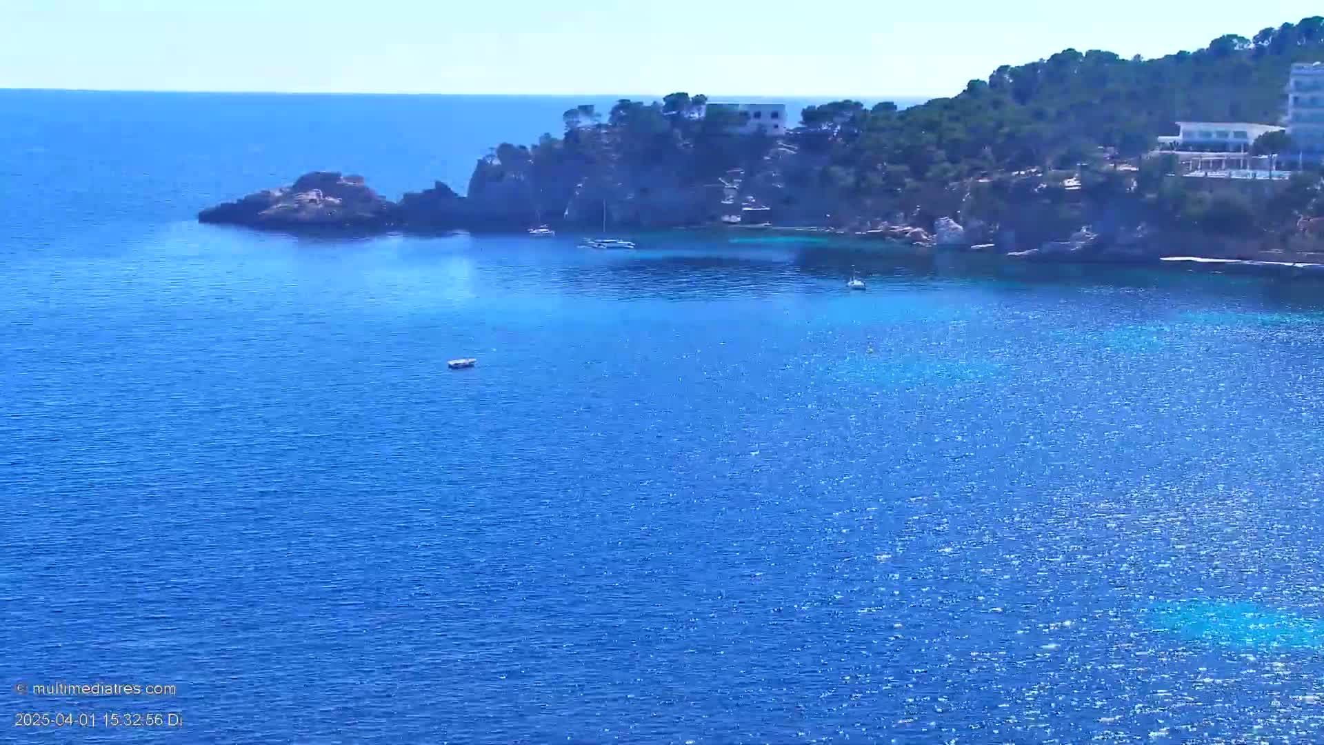 A calm, blue sea laps against a rocky coastline dotted with green vegetation and buildings under a clear, sunny sky.