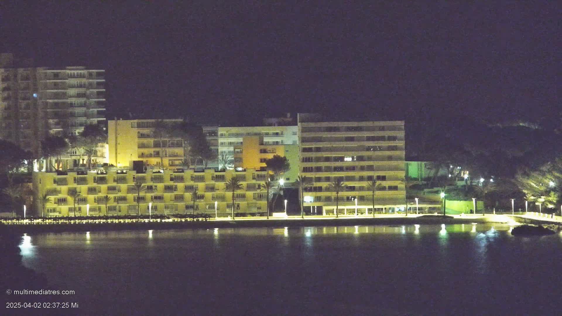 Illuminated multi-story buildings line a waterfront at night, reflected in calm water.