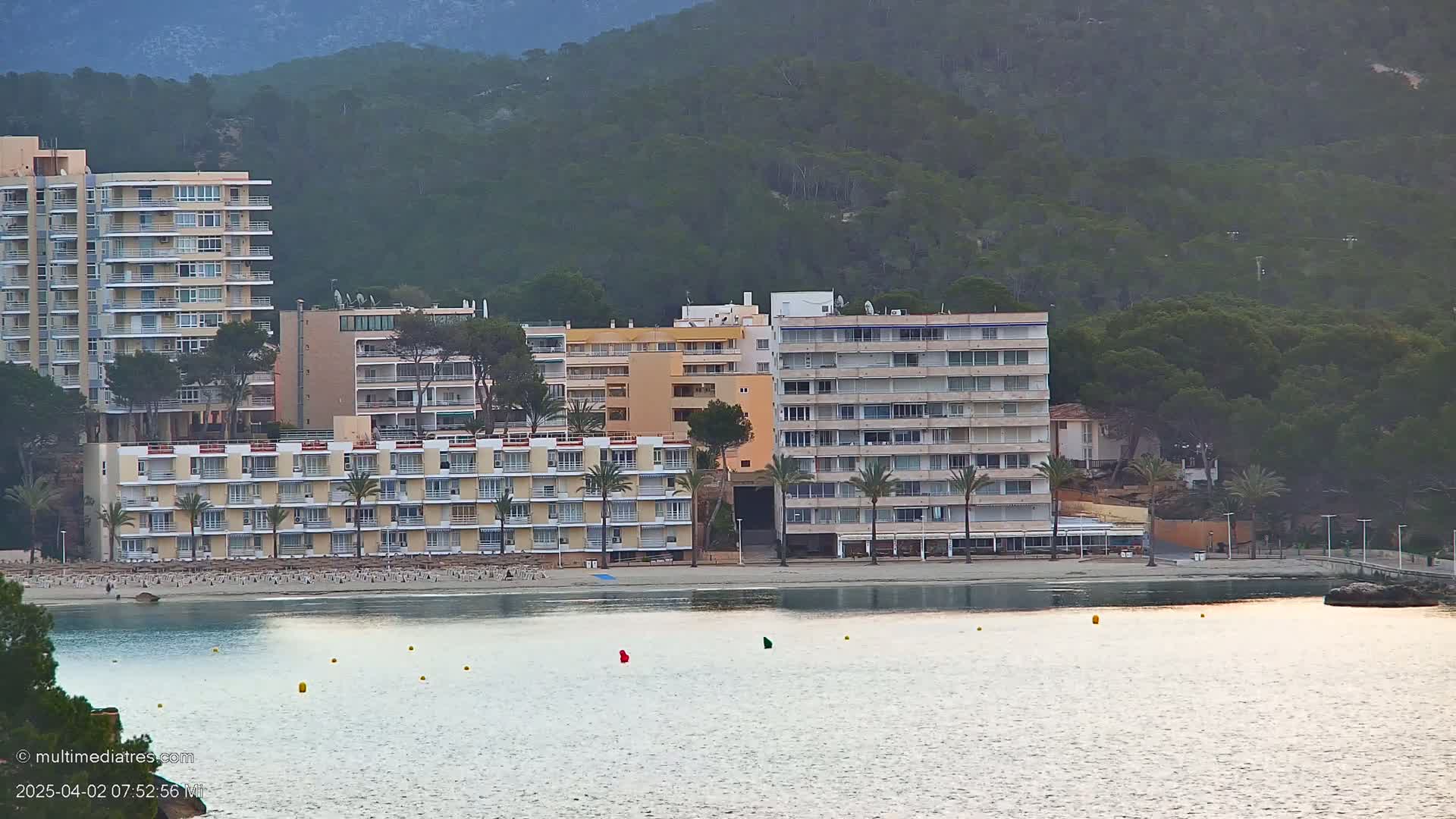 Several multi-story buildings line a sandy beach next to calm water under a mostly cloudy sky.