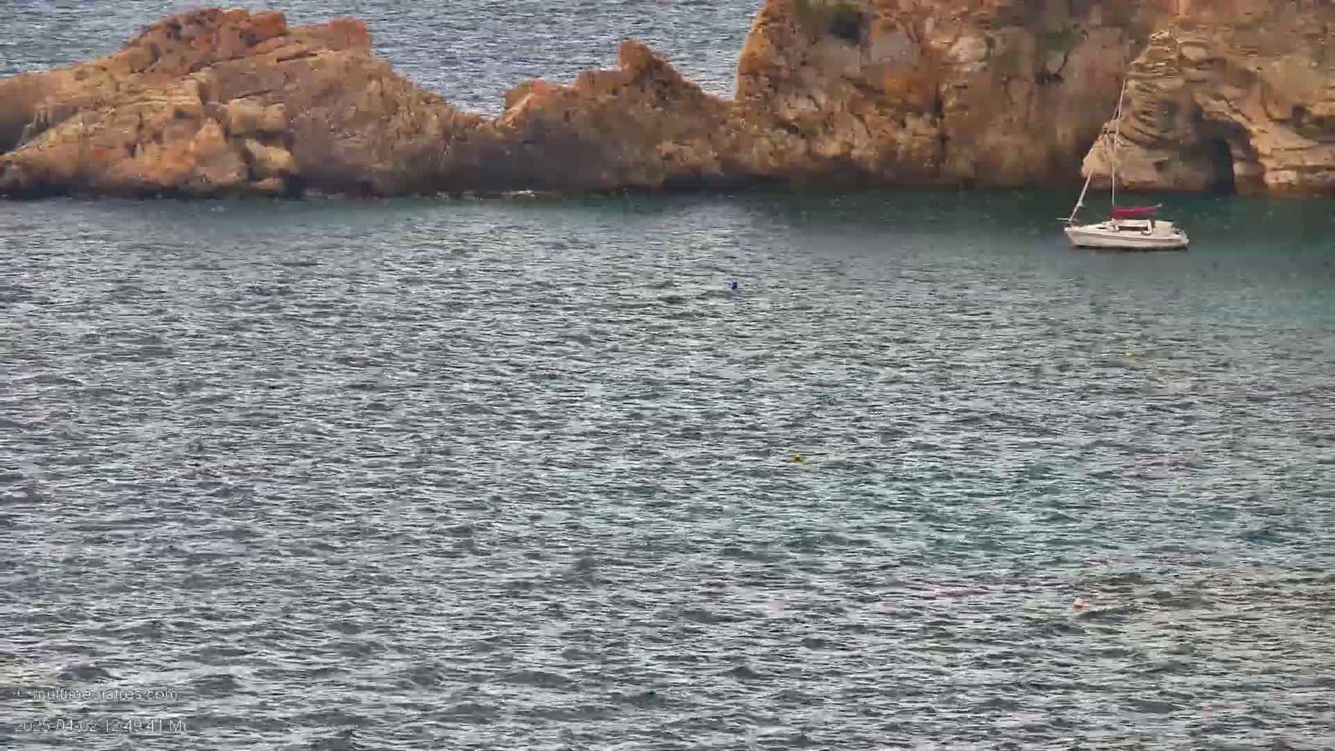 A sailboat is anchored in calm, gray water near a rocky outcrop.