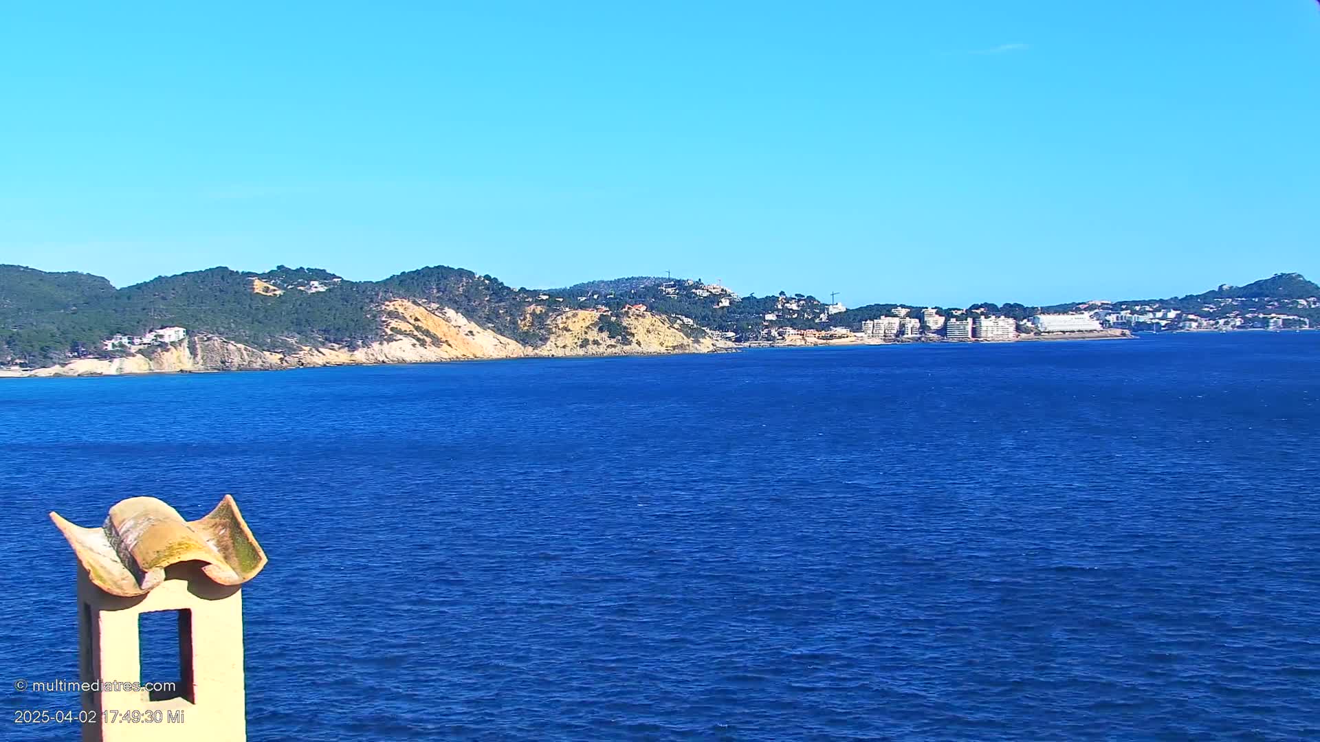 A coastal view shows a calm, deep blue sea extending to a distant shoreline of green hills, tan cliffs, and buildings under a clear blue sky.