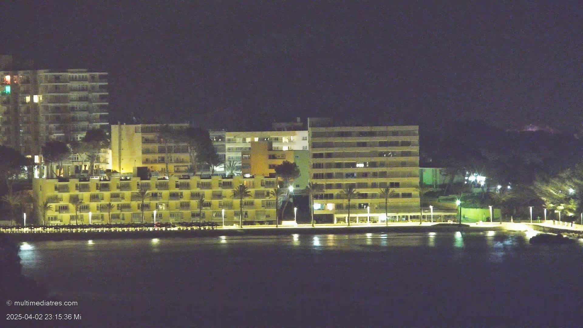 A row of illuminated beachfront buildings stands beside a calm body of water at night.