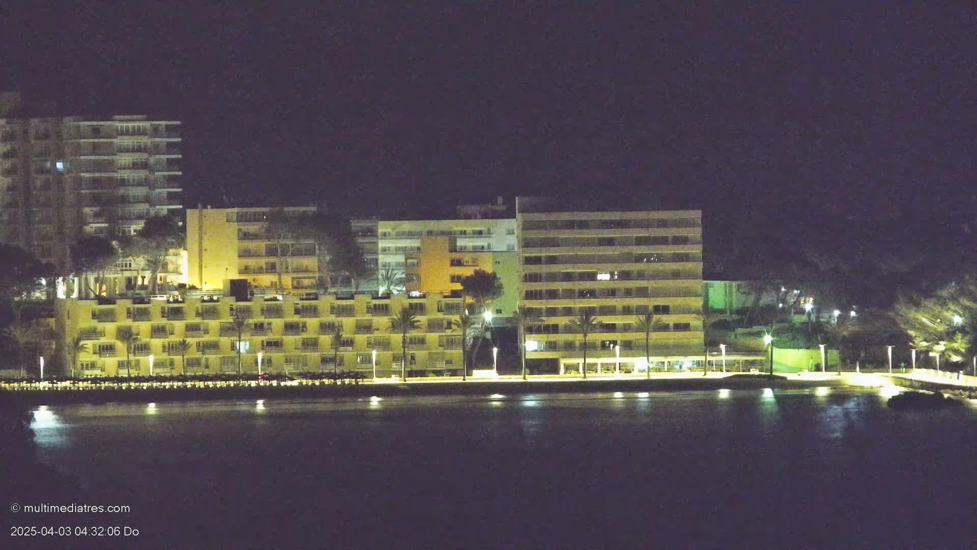 A line of illuminated beachfront hotels and buildings at night, next to calm water.