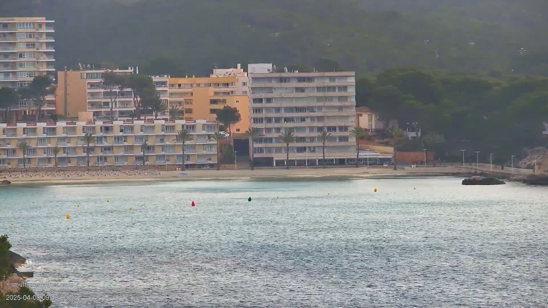 A calm body of water sits in front of several multi-story buildings and a sandy beach, all backed by a hazy, green hill.
