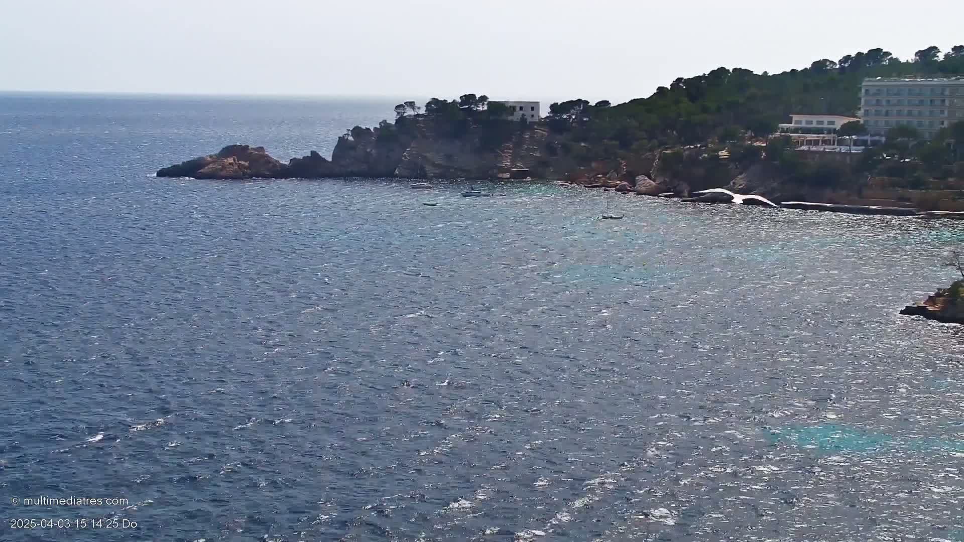 A rocky coastline with a few buildings and some small boats sits on a calm, blue sea under a bright, sunny sky.