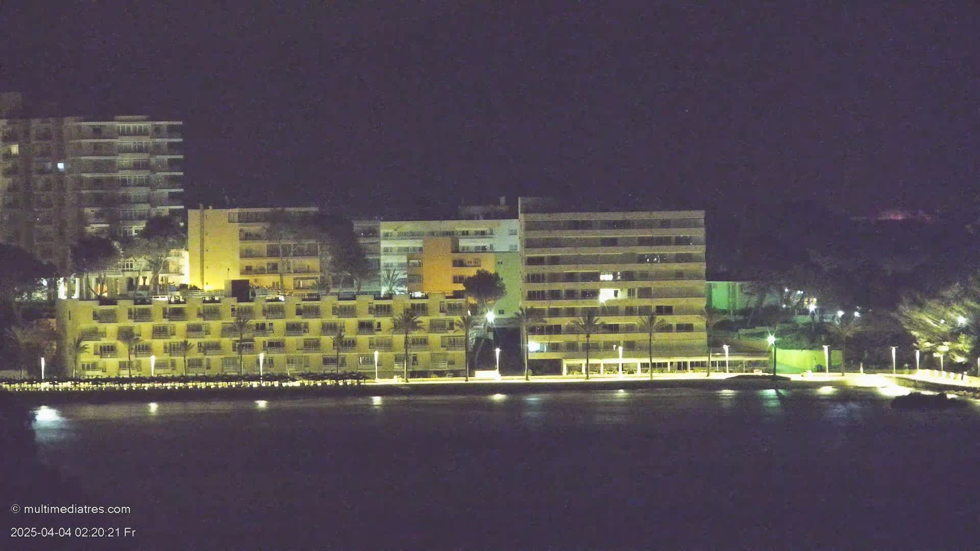 A row of illuminated multi-story hotels sits along a dark, calm body of water at night.