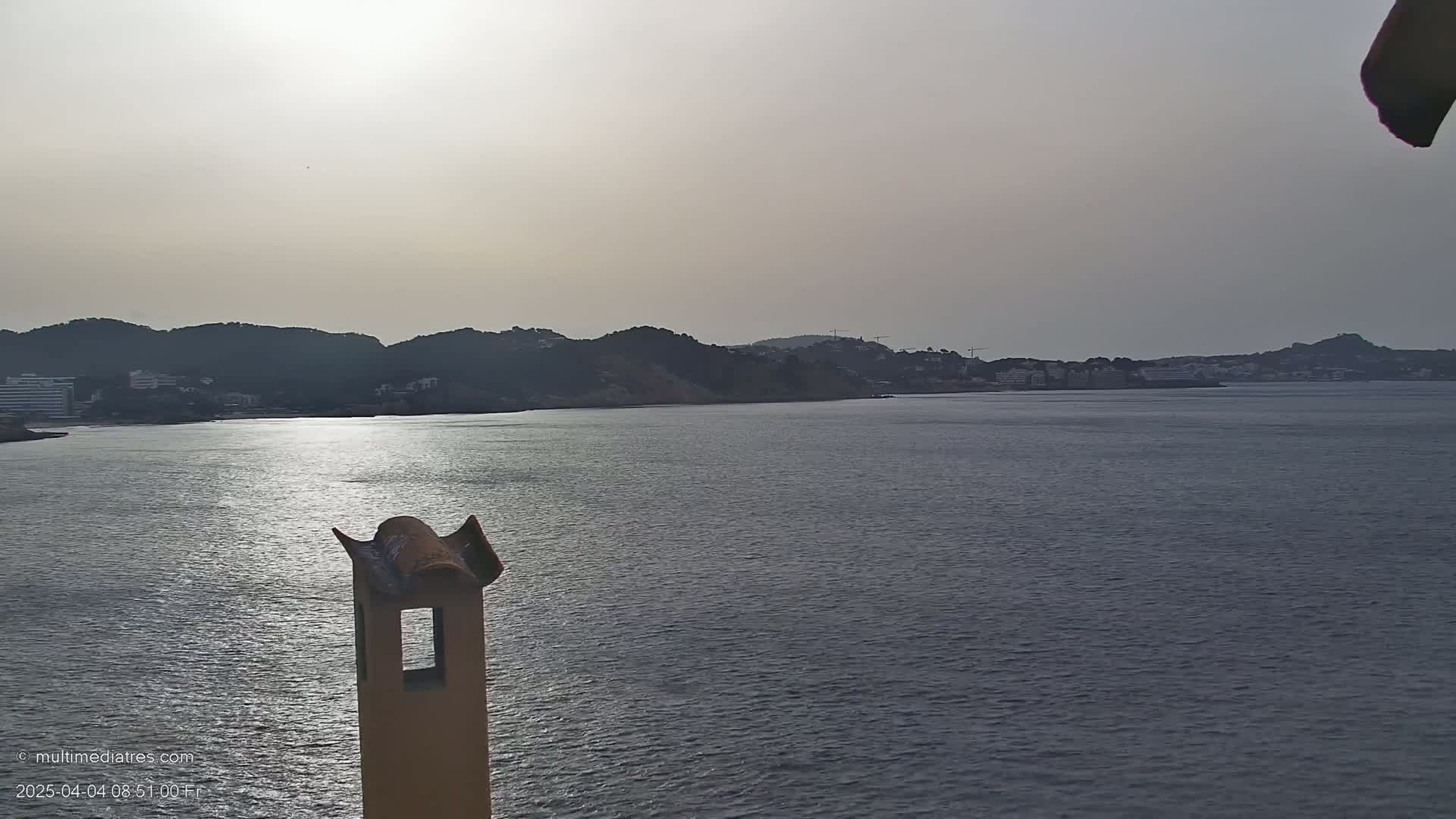 A calm body of water stretches out before a hazy coastline of dark hills and buildings under a bright, overcast sky, partially obscuring a chimney in the foreground.