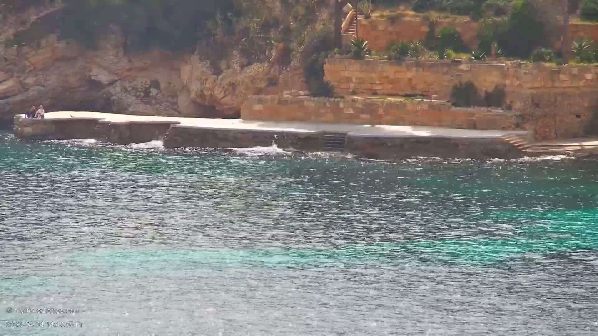 Two people sit on a stone platform overlooking a calm, teal-colored sea next to a rocky, vegetated shoreline.