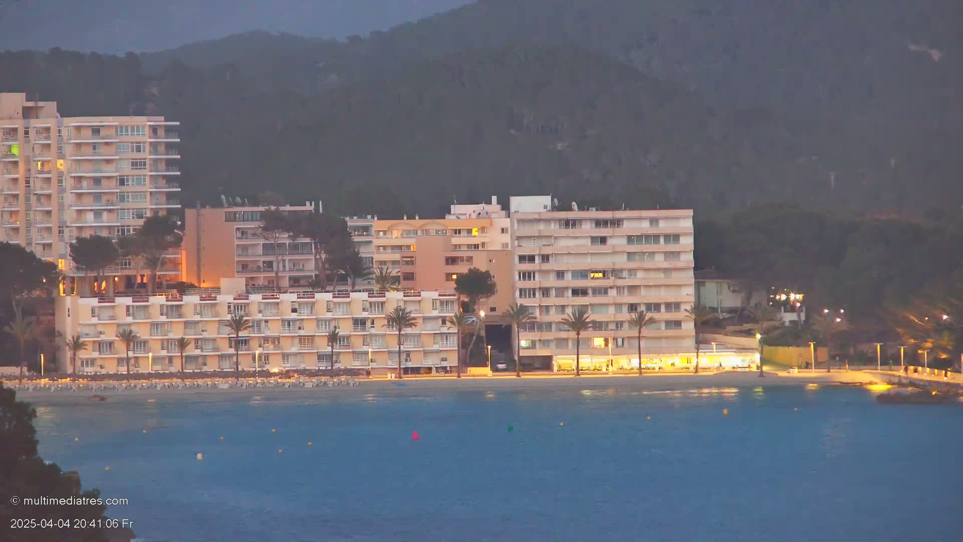 Several multi-story buildings with lights on line a beach at dusk, backed by dark hills.