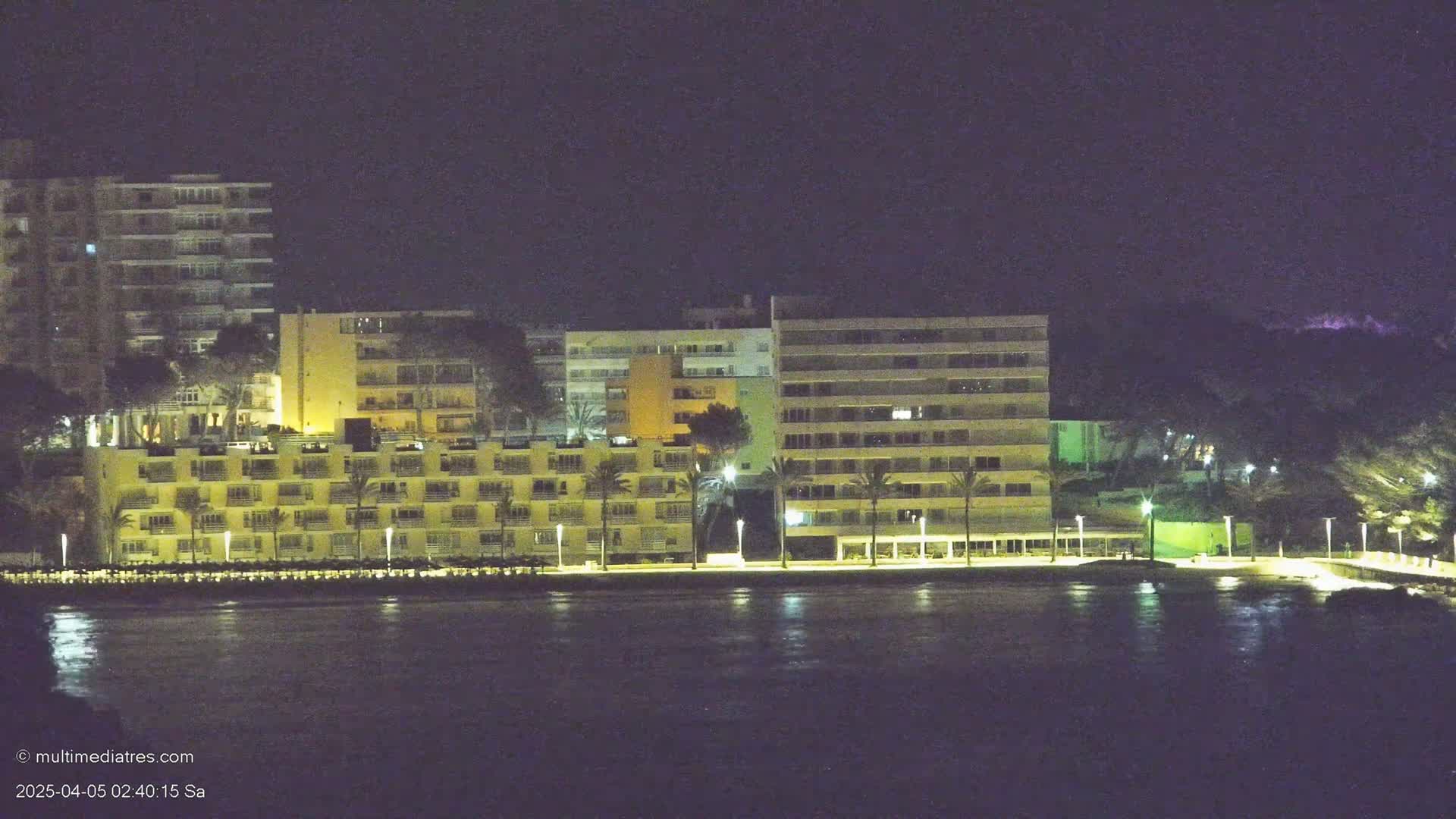 At night, a row of illuminated multi-story buildings lines a dark body of water.