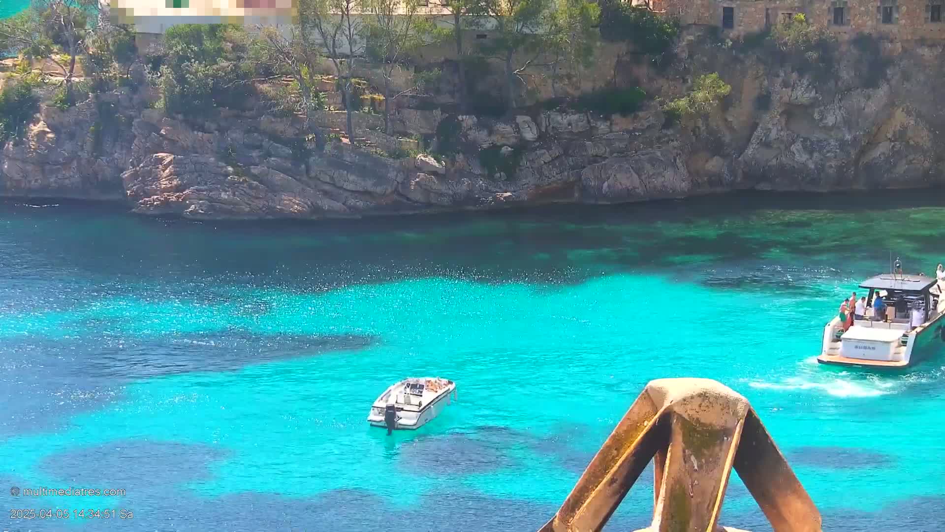 Two boats are on bright turquoise water near a rocky shore with trees and buildings.