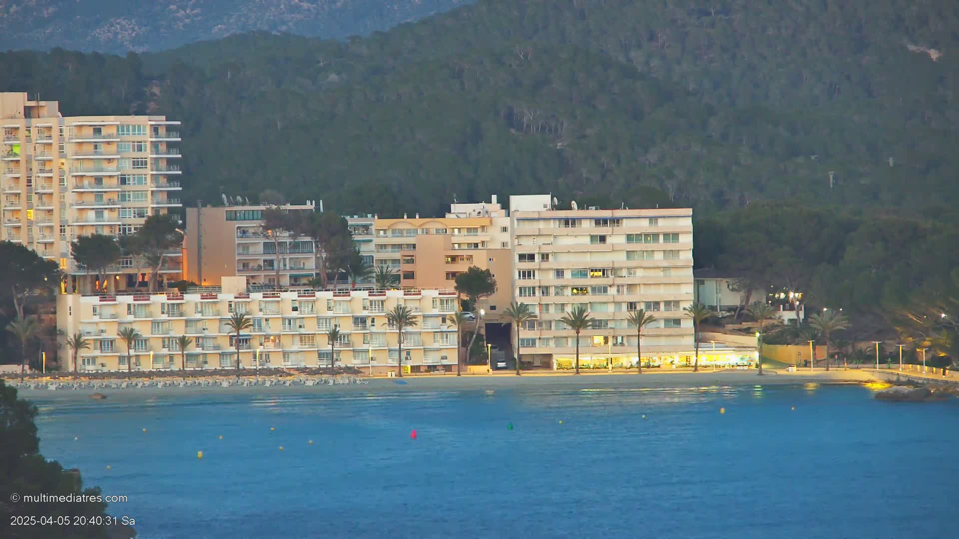 A row of multi-story buildings sits on a beach at dusk, overlooking calm, dark-blue water and a dark-green hillside.