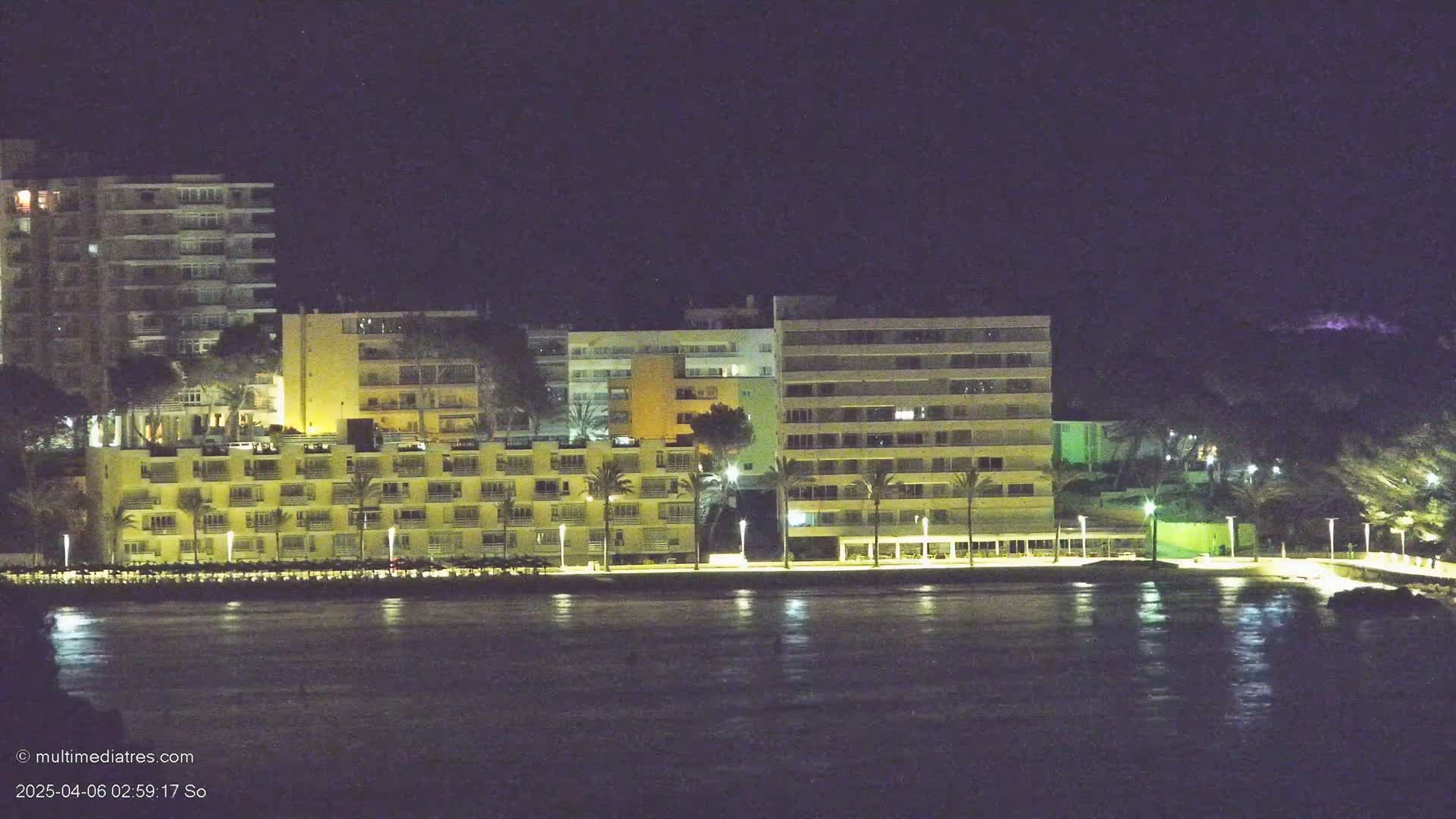 Illuminated beachfront buildings and palm trees are reflected in a dark, still body of water under a night sky.