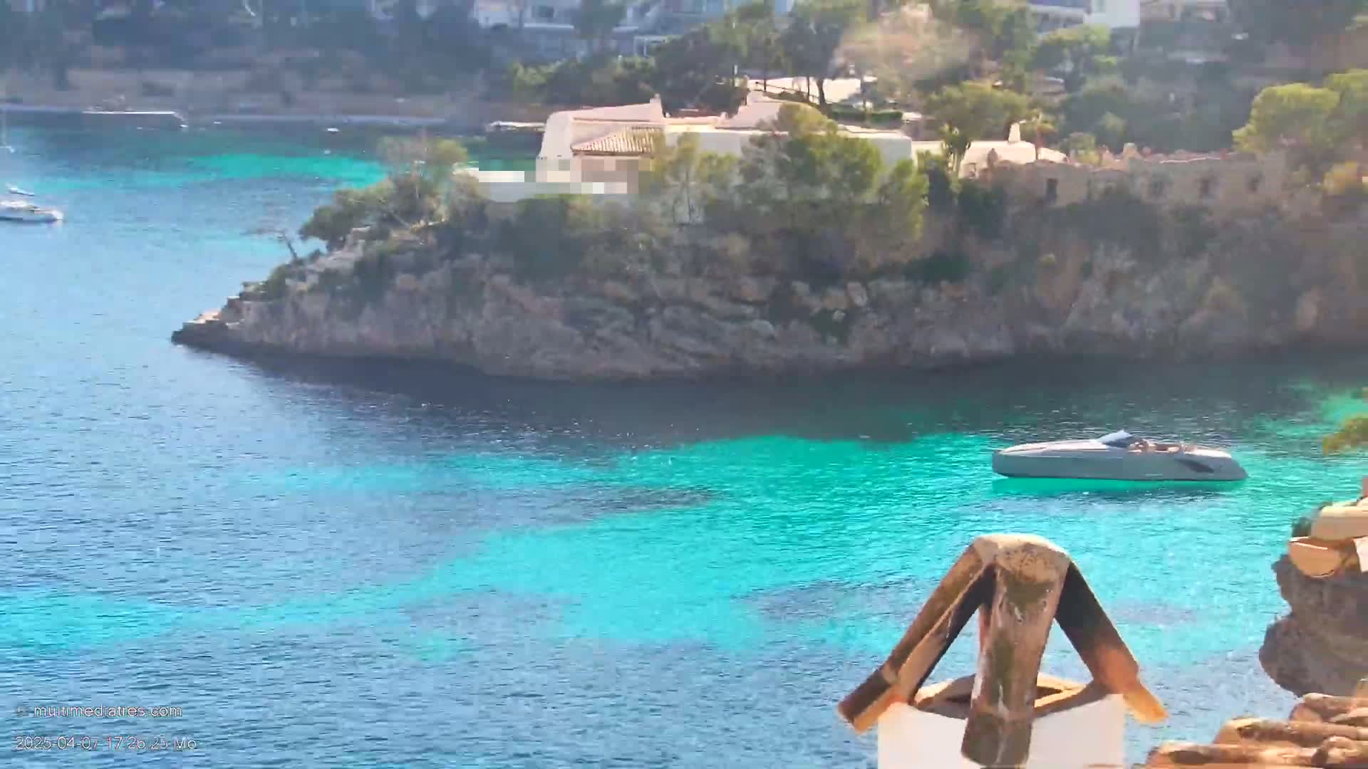 A bright, sunny day shows a rocky cove with turquoise water, a house perched on the cliffs, a lone boat in the water, and other boats visible in the distance.