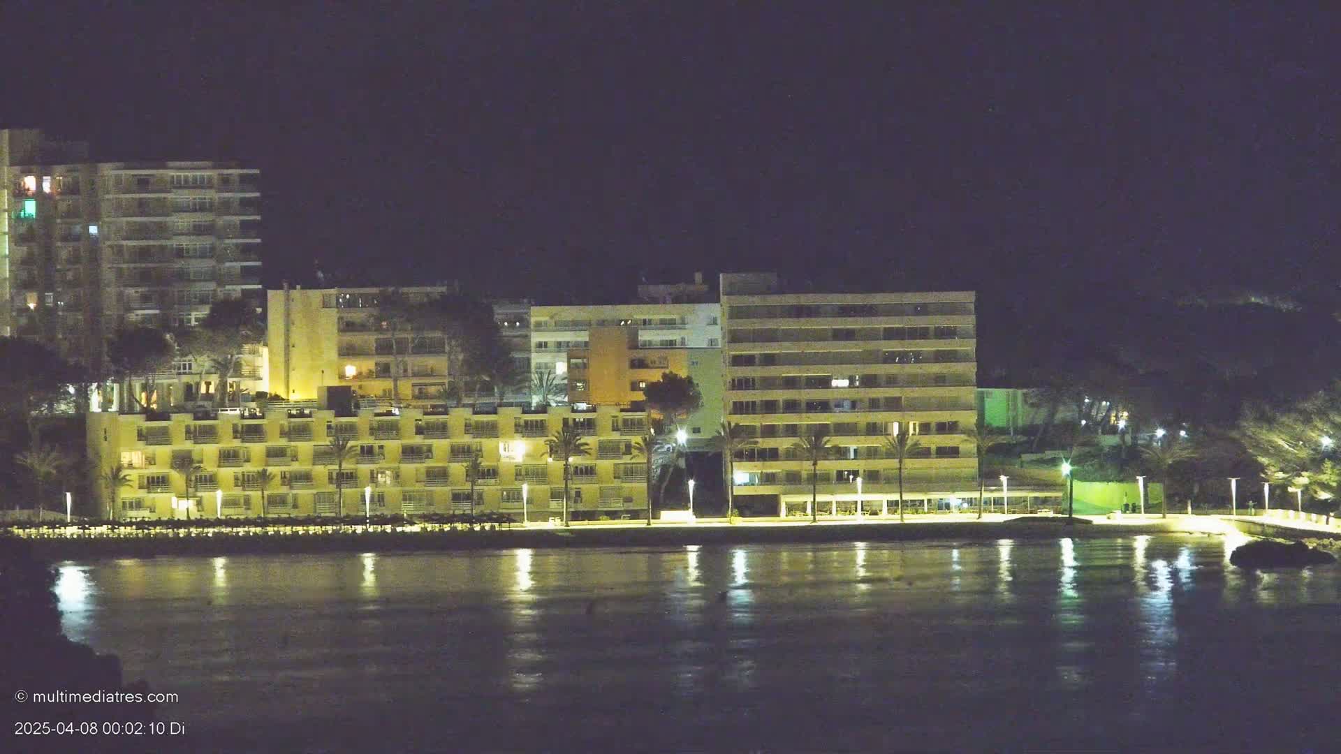 At night, several illuminated multi-story buildings line a waterfront, palm trees dotting the foreground, their reflections shimmering on the calm water.