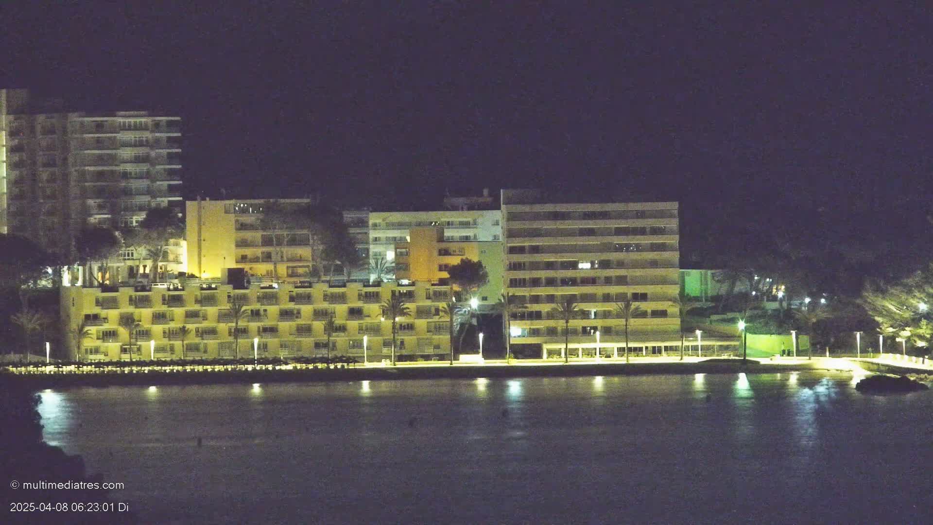 Illuminated multi-story buildings line a waterfront at night, their lights reflecting on calm water.