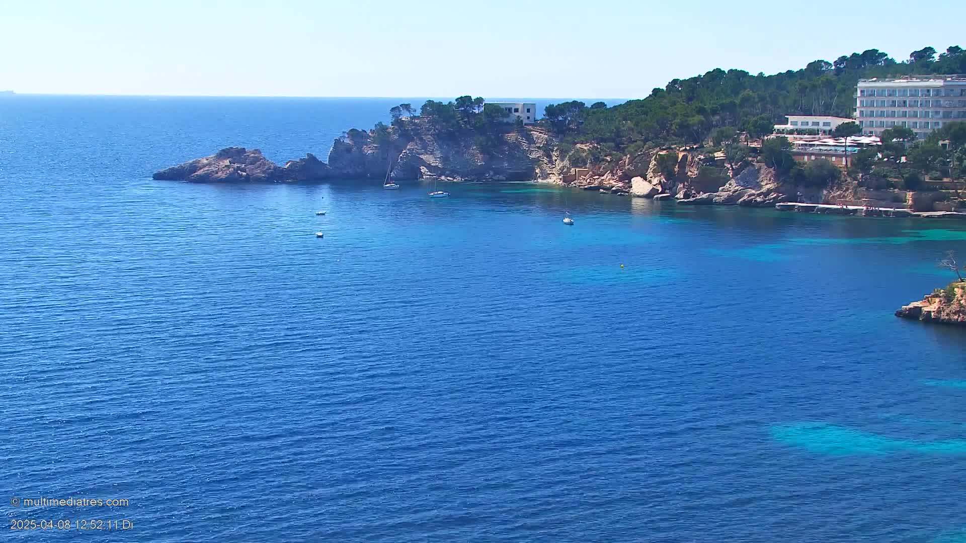 A clear, sunny day shows a rocky coastline with a hotel and several small boats in a calm, blue sea.