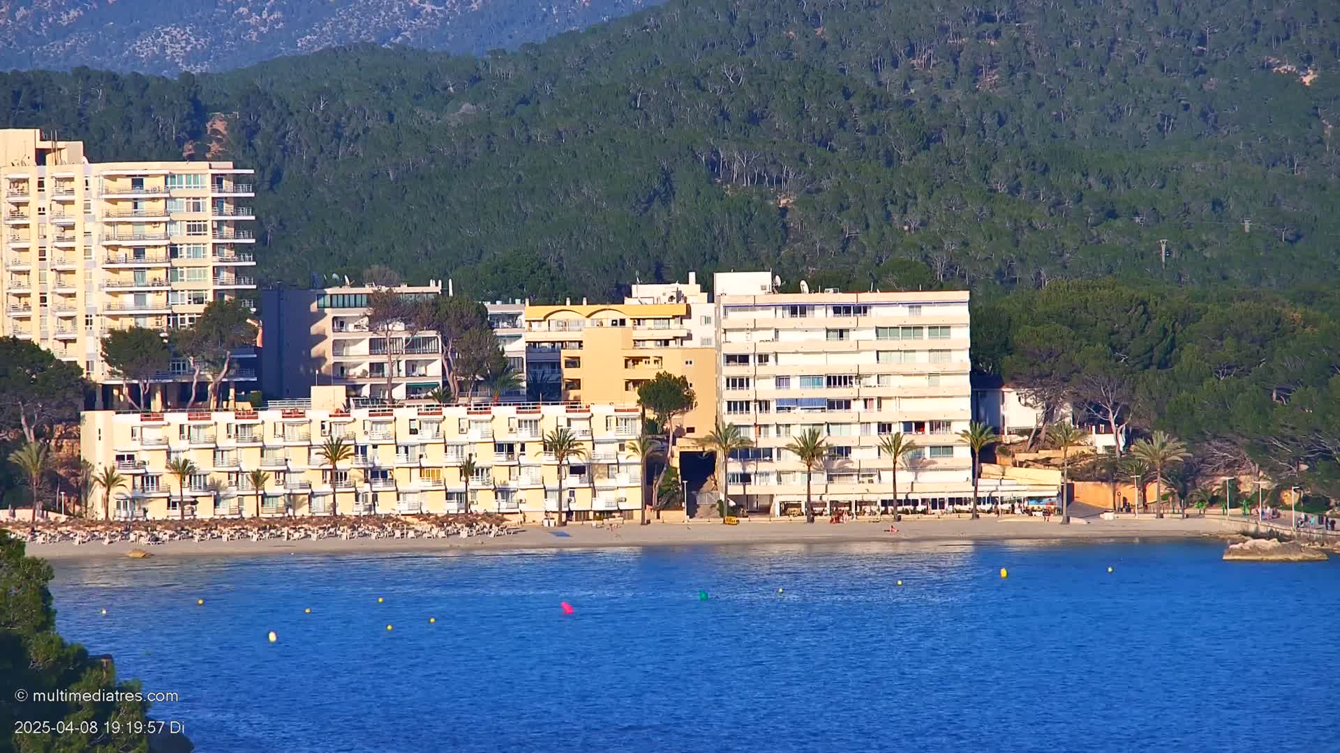 A beachfront scene shows several multi-story hotels situated on a sandy beach next to calm blue water and a forested hillside under a clear sky.